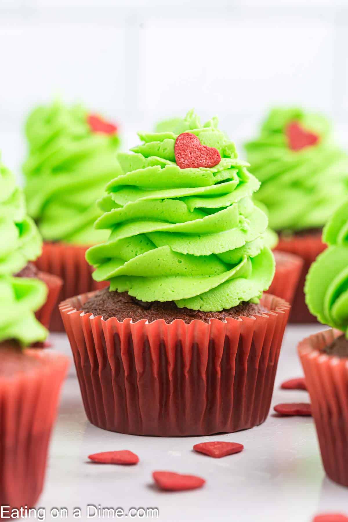 Grinch-inspired chocolate cupcakes with green swirled frosting, topped with red heart sprinkles, capture a mischievously festive vibe. Nestled in red liners and artfully arranged on a white surface, these treats are irresistibly sweet yet delightfully mischief-filled.