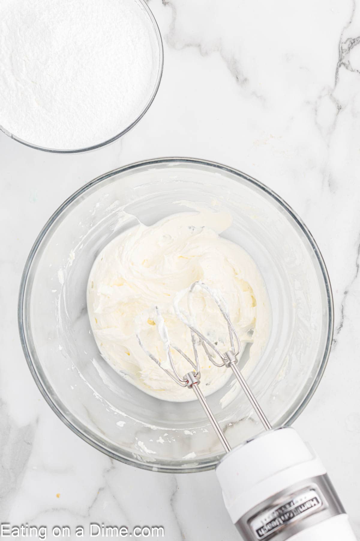 A glass bowl containing creamy whipped mixture is being mixed by an electric hand mixer on a marble countertop, perfect for making Grinch cupcakes. A bowl of powdered sugar is visible nearby.