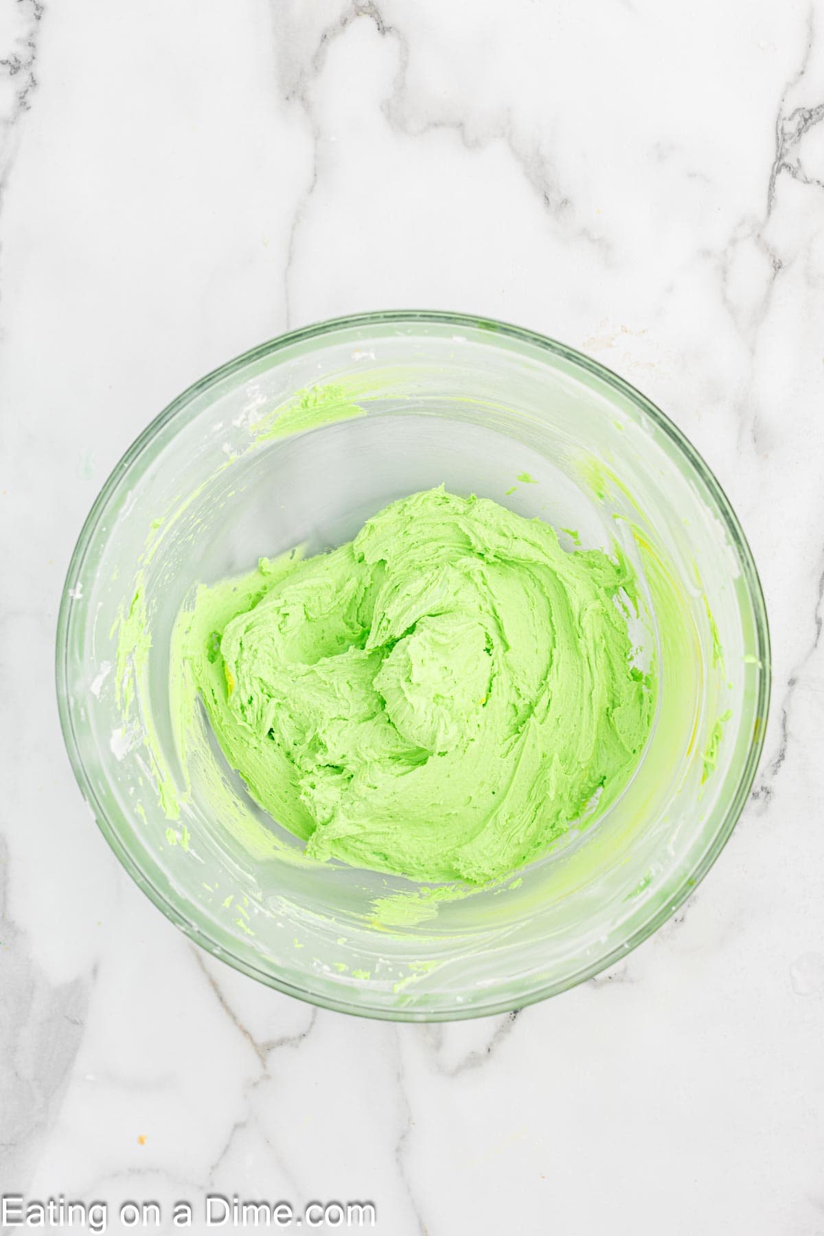 A clear glass bowl containing vibrant green Grinch-inspired frosting sits on a white marble countertop, ready to top festive cupcakes.