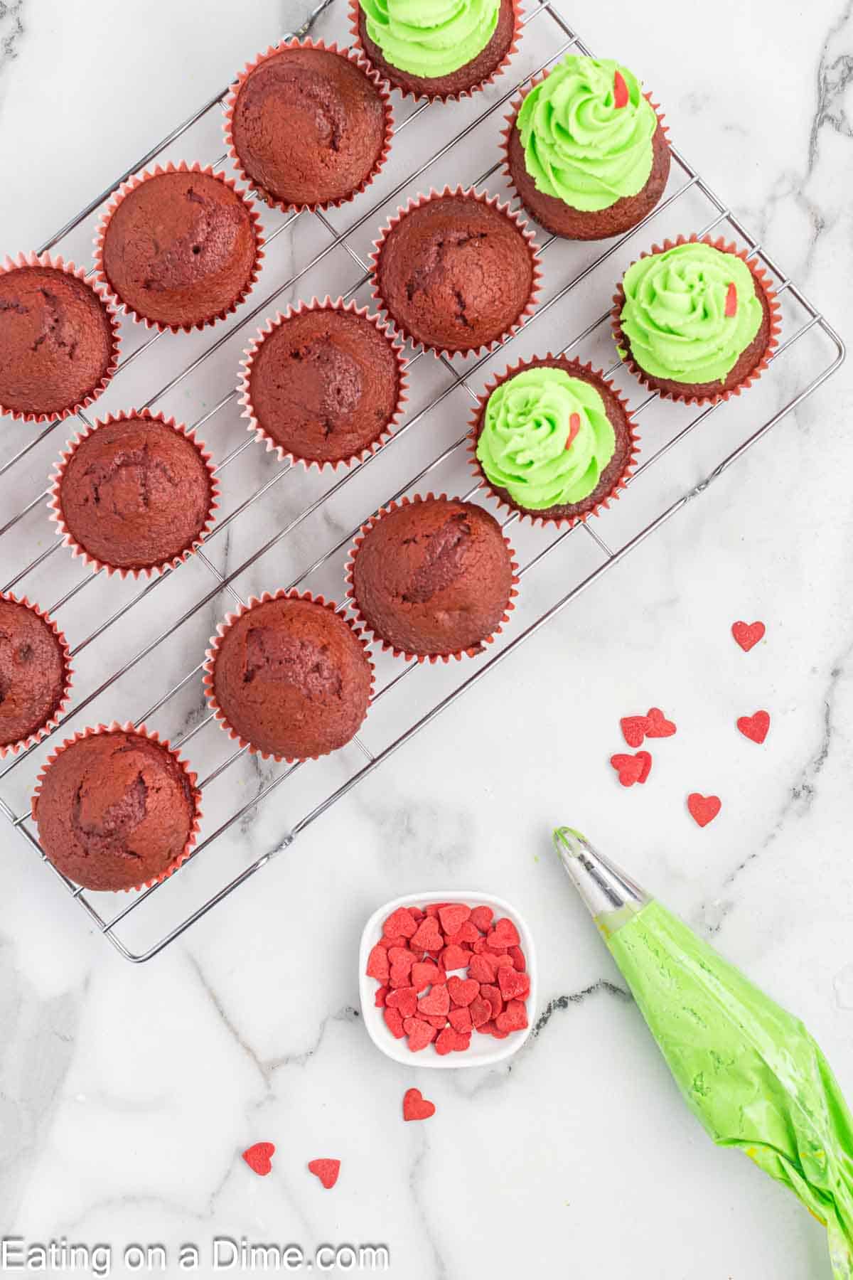 A cooling rack is adorned with Grinch-themed red velvet cupcakes, some topped with green icing and red heart sprinkles. Beside them on the marble countertop, a piping bag filled with green frosting and a small bowl of sprinkles await the next batch of festive treats.