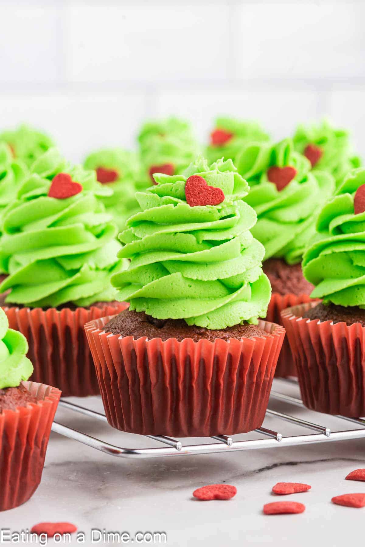 Grinch-themed cupcakes with chocolate bases and red wrappers are topped with bright green frosting, shaped like Christmas trees, and adorned with small red heart sprinkles. They sit cheerfully on a cooling rack, ready to add a whimsical touch to your holiday spread.