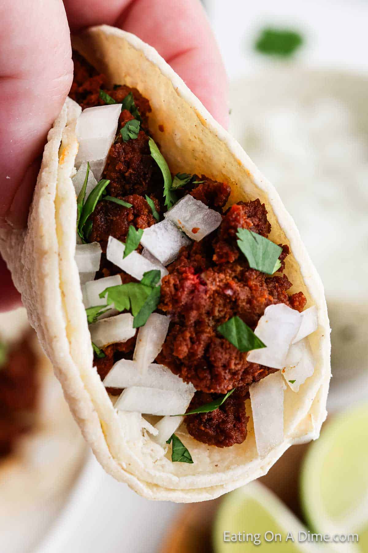 A close-up of a hand holding a street taco filled with crumbled chorizo, chopped onions, and fresh cilantro in a soft tortilla. Lime wedges and green sauce are blurred in the background.