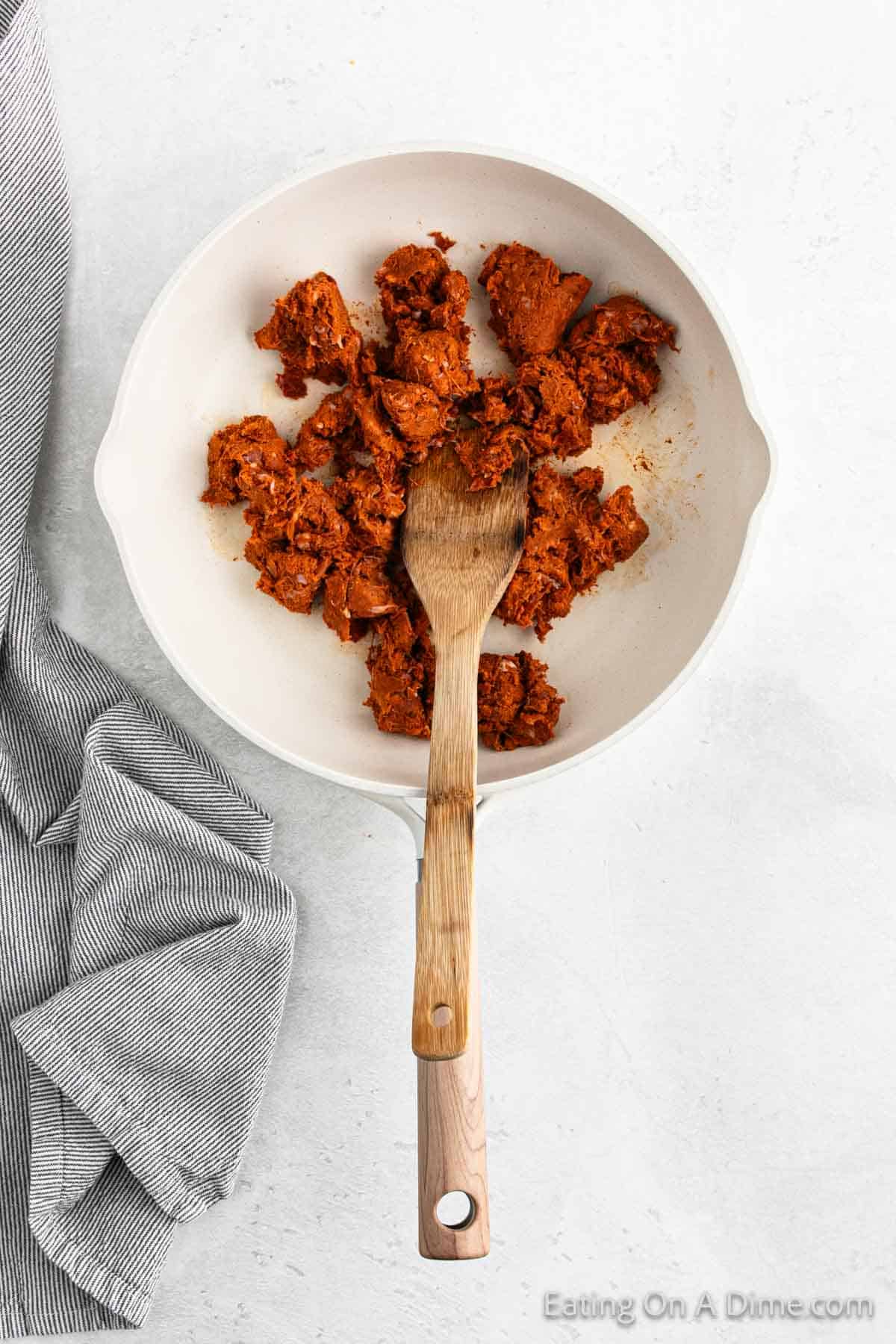 A white pan filled with savory ground beef seasoned like chorizo sits on a light surface. A wooden spatula rests atop this flavorful mix, perfect for street tacos. To the left, a gray and white striped cloth completes the scene.