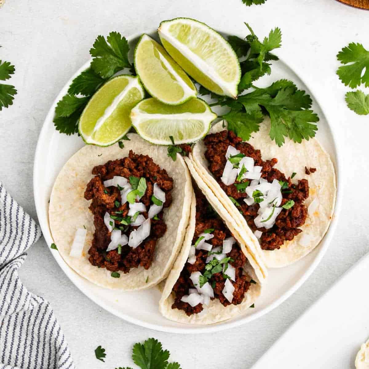 A plate of street tacos, filled with seasoned chorizo, topped with diced onions and cilantro. Lime wedges and cilantro sprigs are arranged on the side. The background features a striped napkin and scattered cilantro leaves.