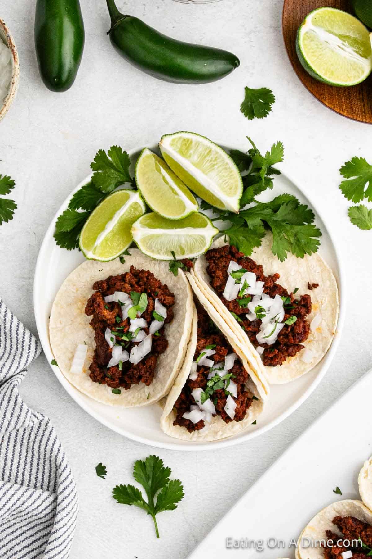 Three street tacos filled with seasoned ground beef and topped with chopped onions and cilantro are arranged on a white plate. Lime wedges and cilantro garnish the dish. A striped napkin and jalapeños are visible in the background, evoking a classic 3-ingredient simplicity.