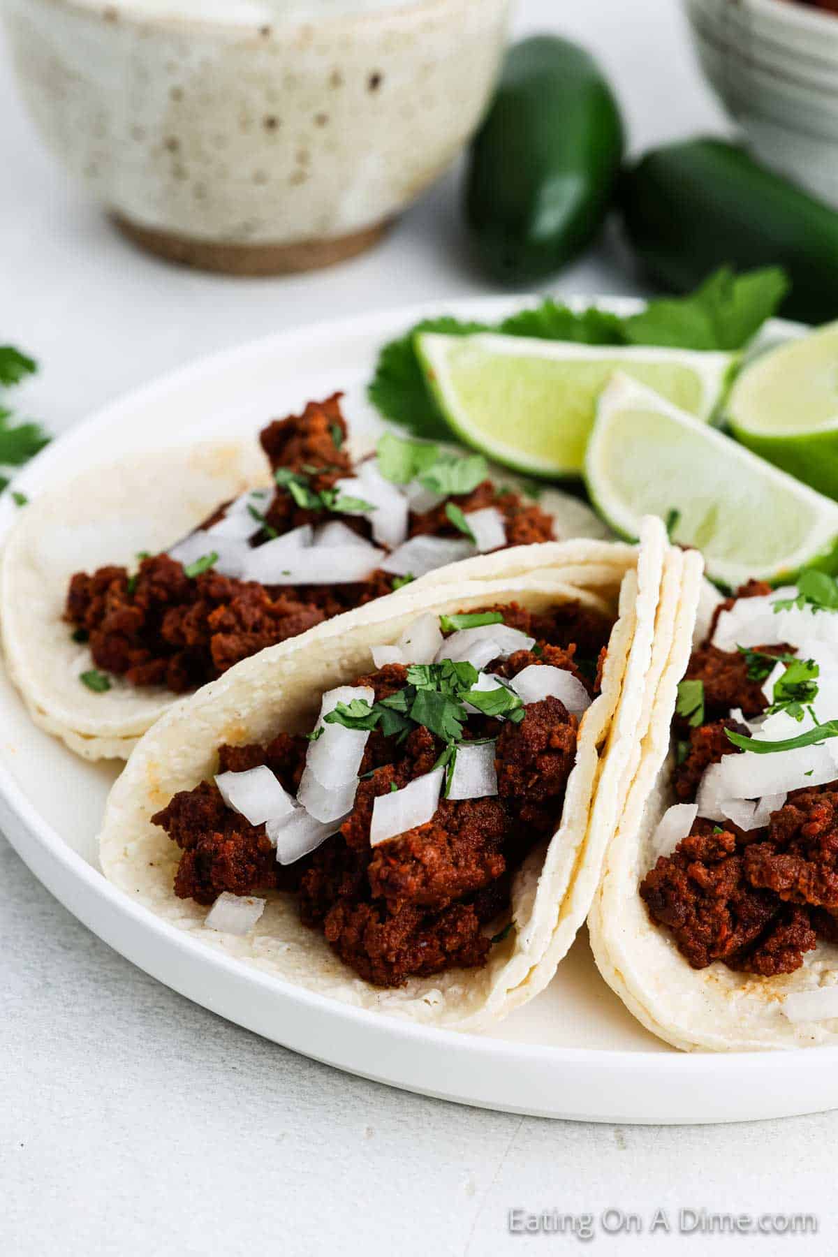 A plate of mouth-watering street tacos filled with seasoned chorizo, topped with chopped onions and cilantro. Lime wedges and jalapeños are on the side, adding zest to this 3-ingredient delight. A bowl is partially visible in the background.