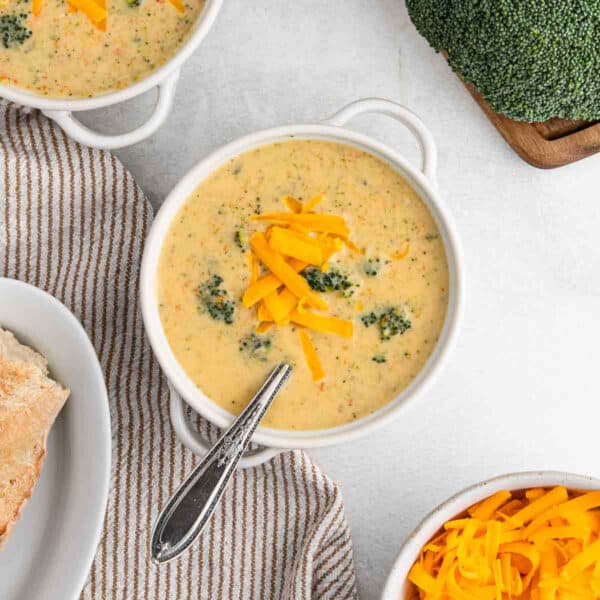 A bowl of creamy broccoli cheddar soup topped with extra cheese rests on a striped cloth. A spoon sits in the bowl, accompanied by a bread slice on a plate and more shredded cheese nearby. Fresh broccoli peeks from the corner, completing this cheesy delight.