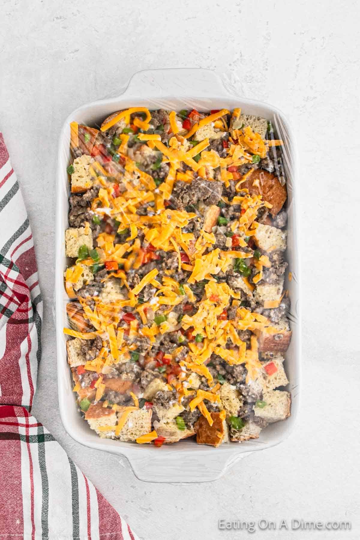 Casserole dish with shredded cheese, cooked sausage, red bell pepper, green bell pepper and diced bread covered with plastic wrap 