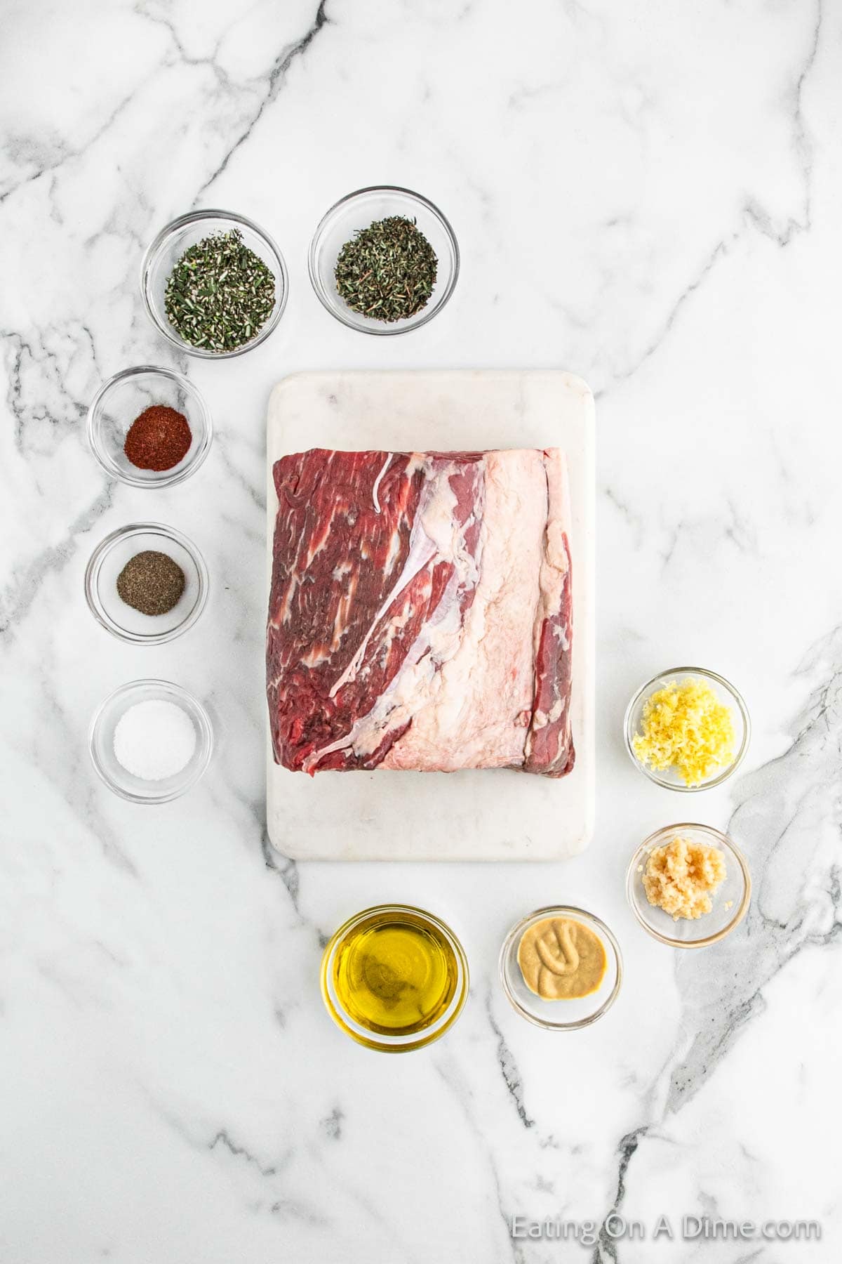 A marble countertop showcases a large cut of raw beef, perfect for a Christmas roast, on a white cutting board. It's surrounded by small bowls filled with herbs, spices, olive oil, mustard, minced garlic, and crushed herbs—ready for seasoning.