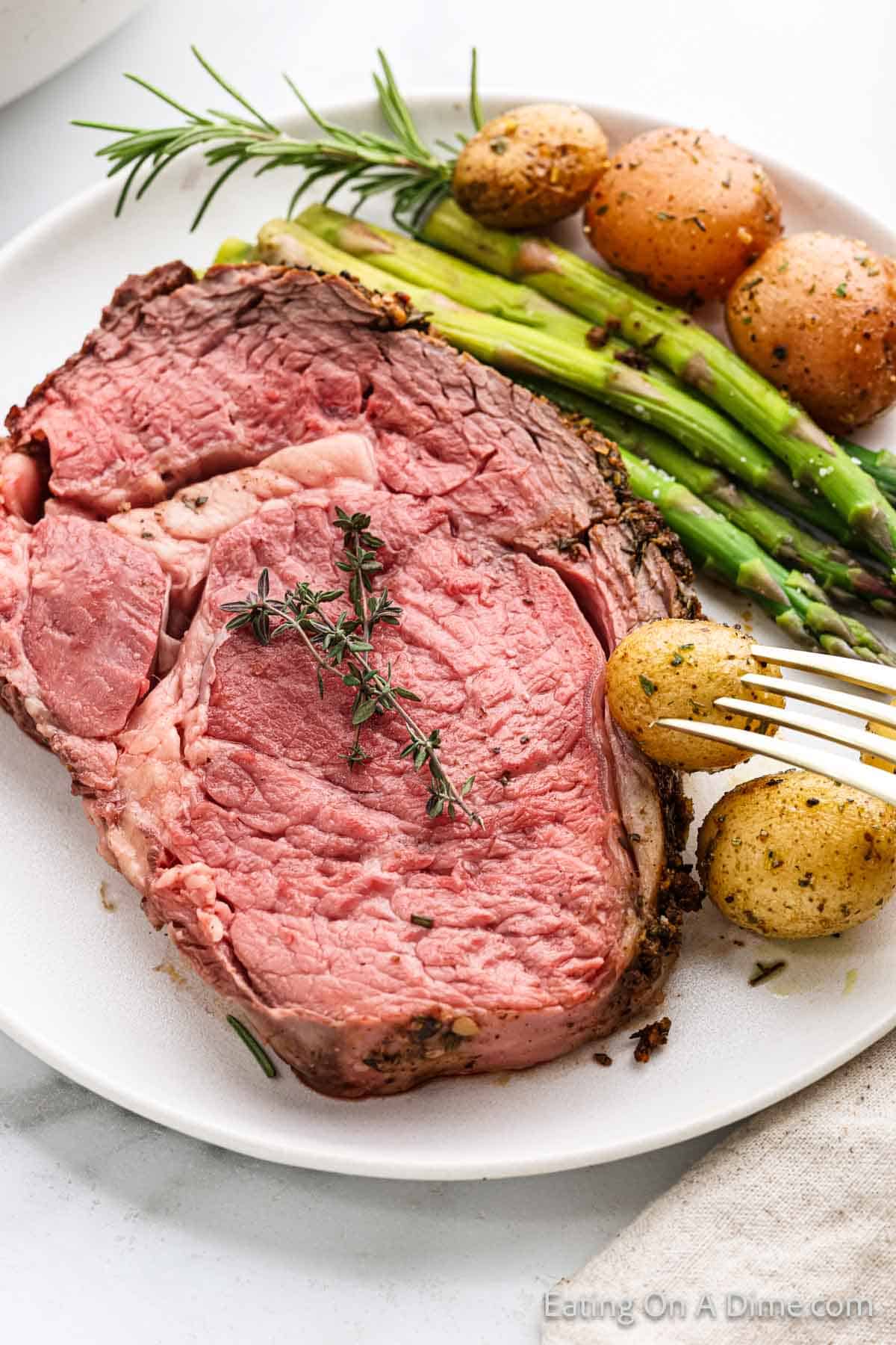 A plate with a succulent slice of rare roast beef garnished with herbs is reminiscent of Christmas cheer. Accompanied by roasted baby potatoes and asparagus spears, this festive roast is completed with a fork resting beside the potatoes.