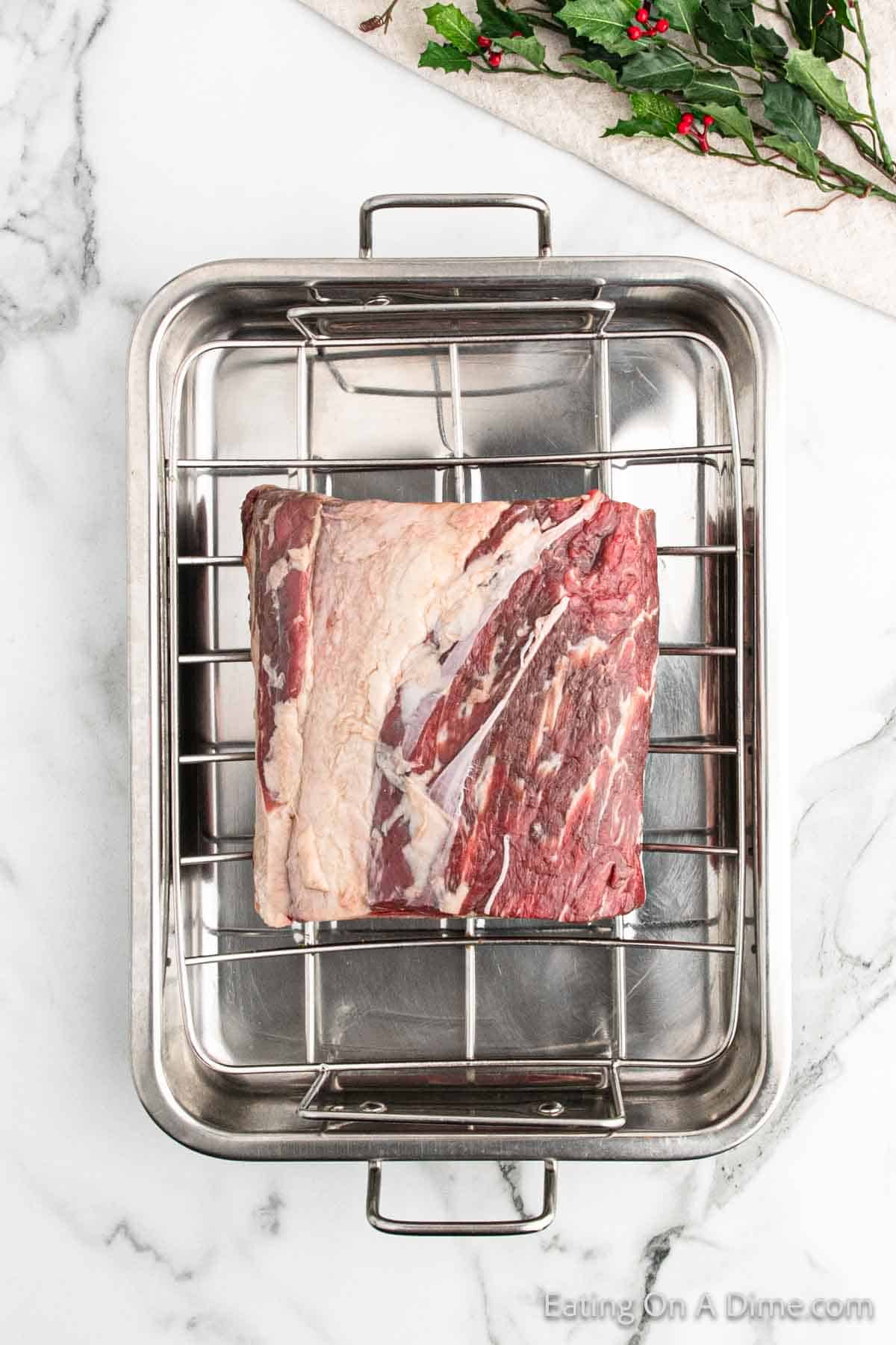 A raw beef roast rests on a metal rack inside a roasting pan atop a white marble countertop, ready to become the centerpiece of your Christmas feast. In the top right corner, festive sprigs of green holly with red berries add holiday cheer.