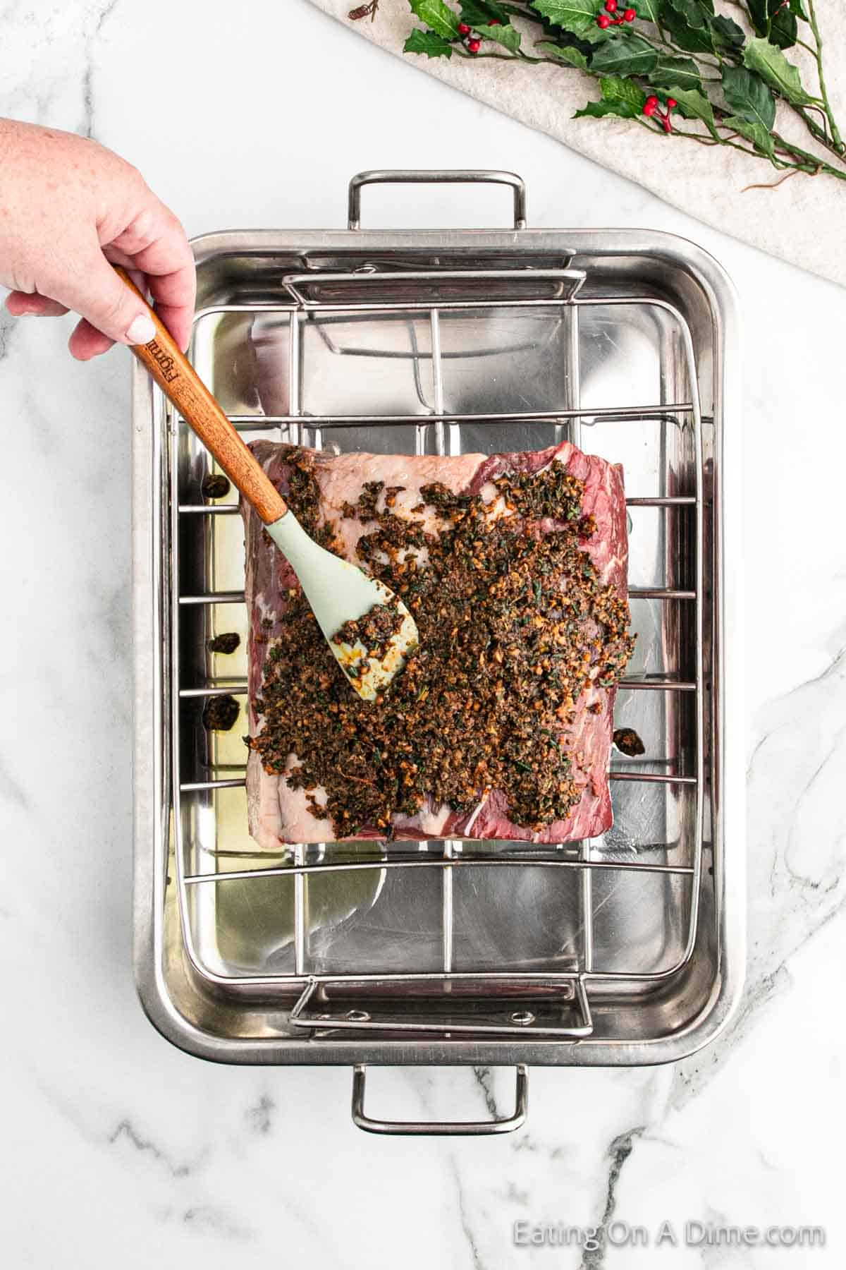 A hand spreads a dark herb mixture onto a raw beef roast in a metal roasting pan with a wire rack, capturing the essence of Christmas preparation. The pan rests on a white marble surface, with a festive sprig of holly adorning the corner.