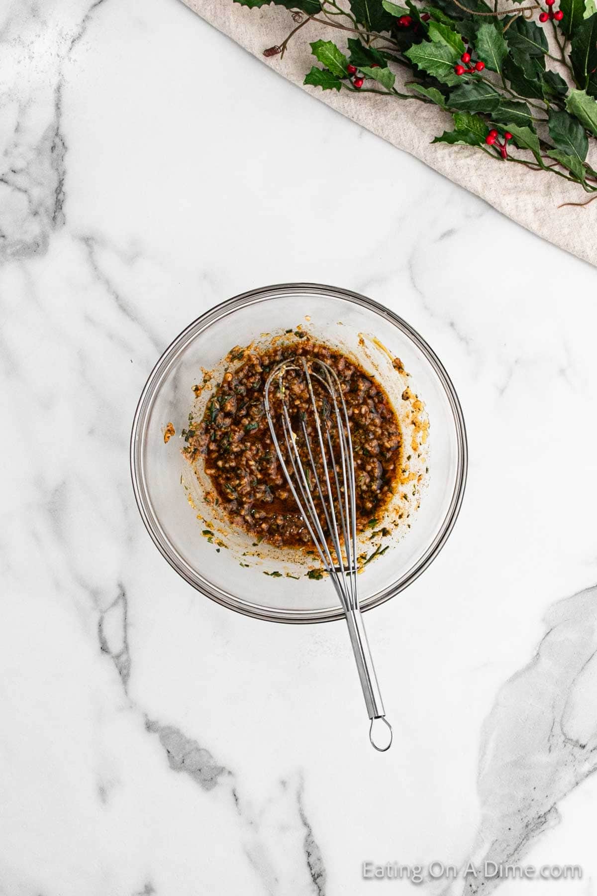 A glass bowl filled with a mixed herb marinade and a whisk sits on the marble countertop, ready to complement your Christmas roast. Fresh holly branches with red berries elegantly adorn the top edge of the image, adding a festive touch.