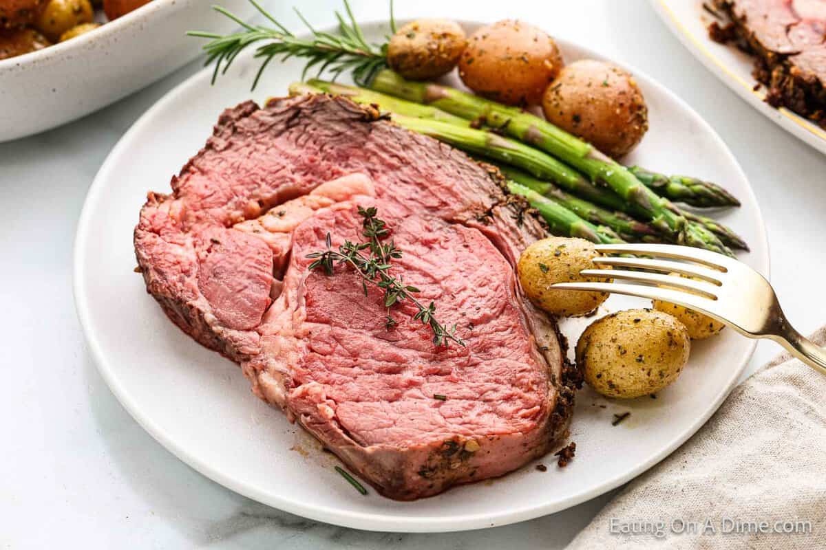 A plate with a thick slice of pink, juicy prime rib roast garnished with fresh thyme evokes the festive spirit. Accompanied by roasted baby potatoes and bright green asparagus spears, it's a Christmas feast on your plate. A fork is set next to the potatoes.