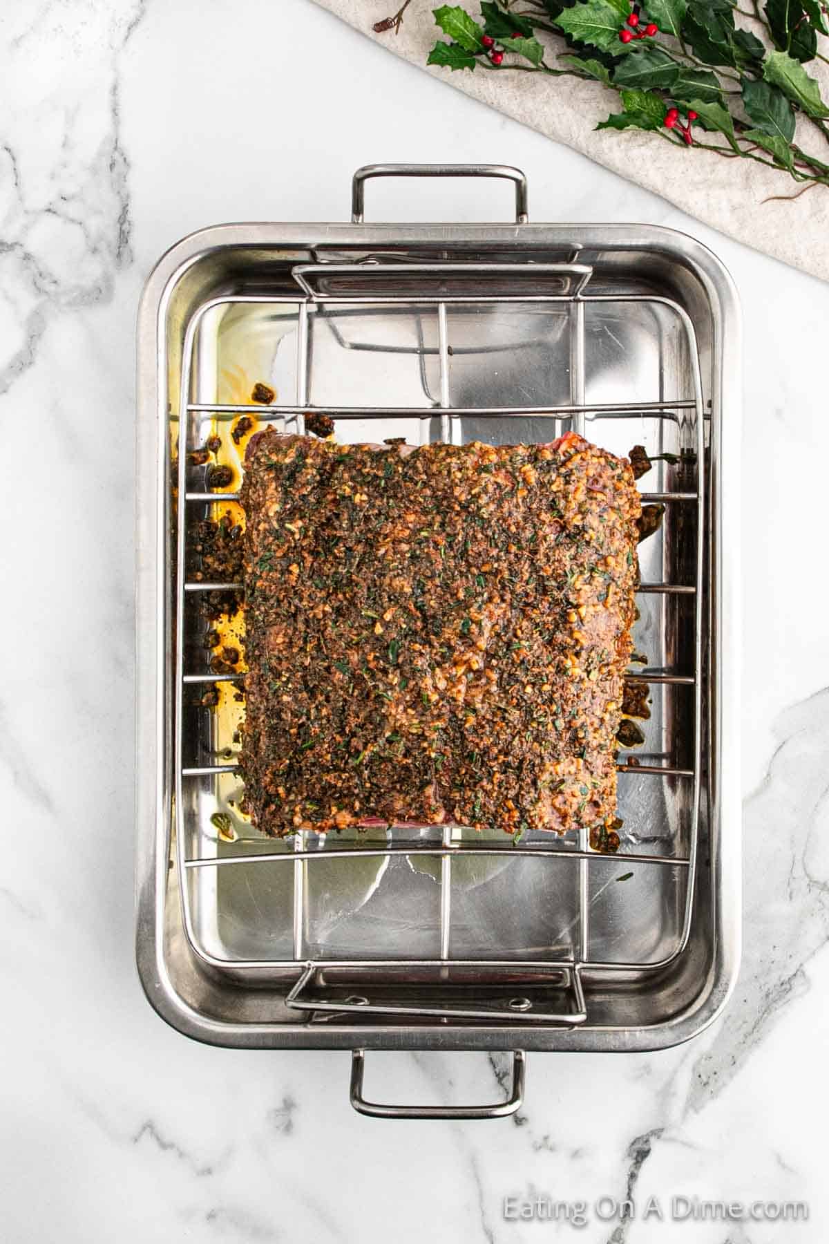 A Christmas holiday roast coated with savory herbs and spices sits on a roasting rack inside a metal pan. The pan is placed on a marble countertop, and a festive sprig of holly decorates the top right corner.