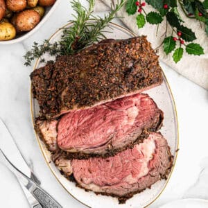 Sliced herb-crusted roast beef on an oval platter, garnished with fresh rosemary and thyme. A bowl of roasted potatoes and a festive Christmas holly decoration add to the warm, seasonal ambiance in the background.