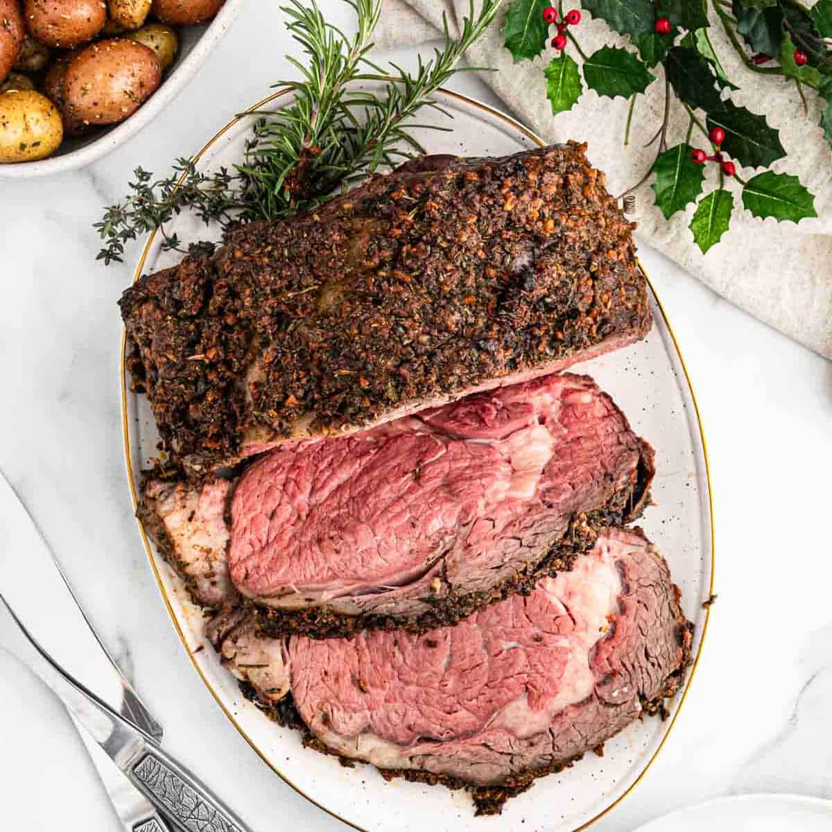 A roasted prime rib with a crispy herb crust is sliced on a platter, perfect for a festive Christmas feast. Garnished with fresh rosemary, it's served alongside a bowl of roasted potatoes and a decorative sprig of holly. Silverware sits elegantly beside the plate on a marble surface.