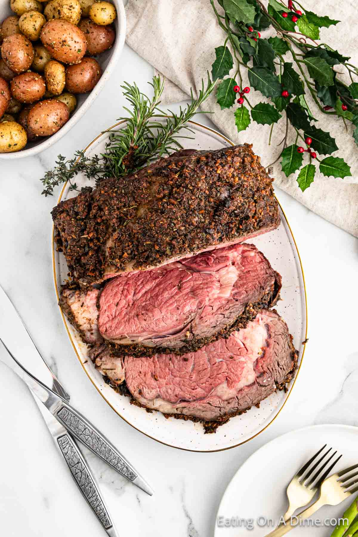 A platter with slices of Christmas roast beef garnished with herbs, accompanied by a bowl of roasted potatoes. Silver utensils are placed nearby on a white tablecloth. Decorative holly and a sprig of rosemary add a festive touch.