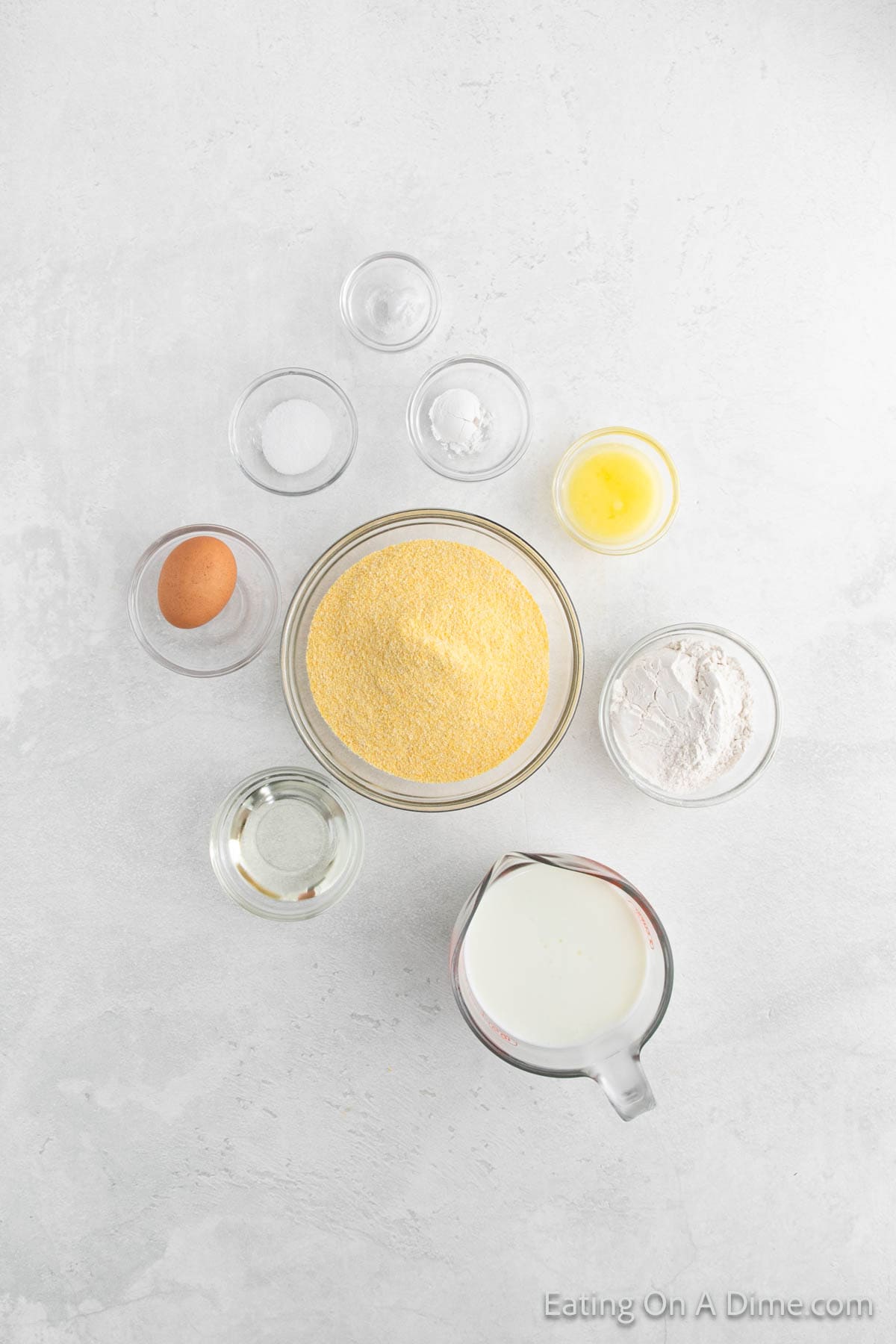 Top view of cornbread ingredients for a delicious fried cornbread recipe on a light surface, including cornmeal, milk, an egg, flour, melted butter, oil, baking powder, sugar, and salt—each perfectly portioned in separate bowls or a measuring cup.