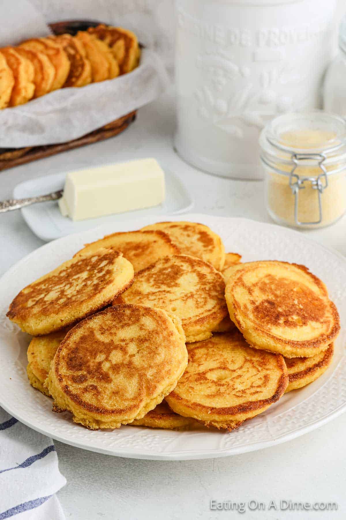 A plate of golden brown hoecakes, reminiscent of a classic fried cornbread recipe, stacked neatly. In the background, there's a basket with more hoecakes, a stick of butter on a dish, a jar of cornmeal, and a white container on a light-colored table.