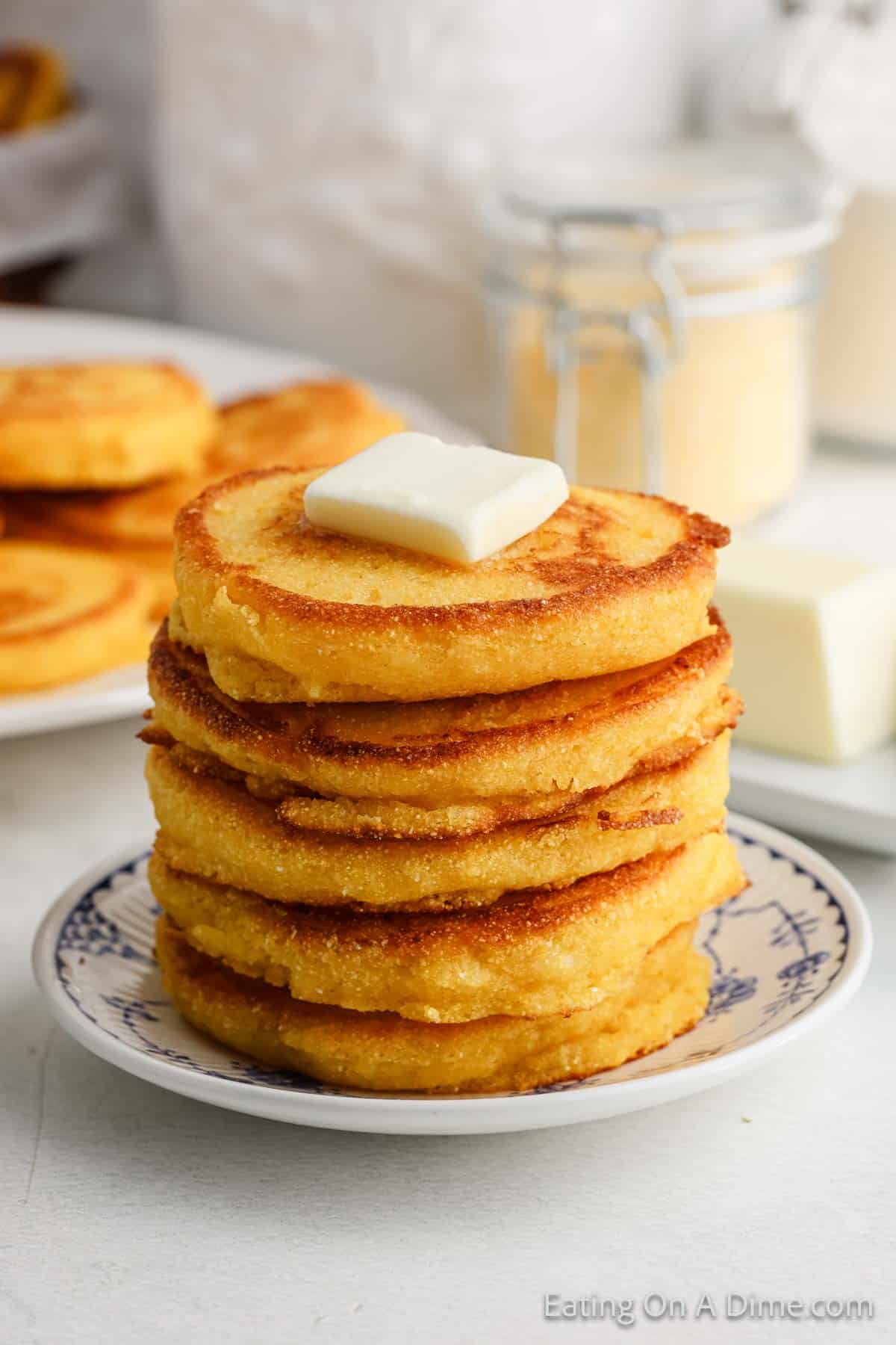 A stack of golden brown hoe cakes, reminiscent of a classic fried cornbread recipe, is topped with a pat of butter on a decorative plate. In the background, a slab of butter and a jar of cornmeal hint at the cornbread recipe’s delicious foundation.