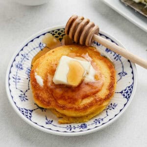 A stack of pancakes topped with a pat of butter and drizzled with honey sits on a decorative plate, reminiscent of classic fried cornbread. A wooden honey dipper rests on the edge, ready to add a touch more sweetness.