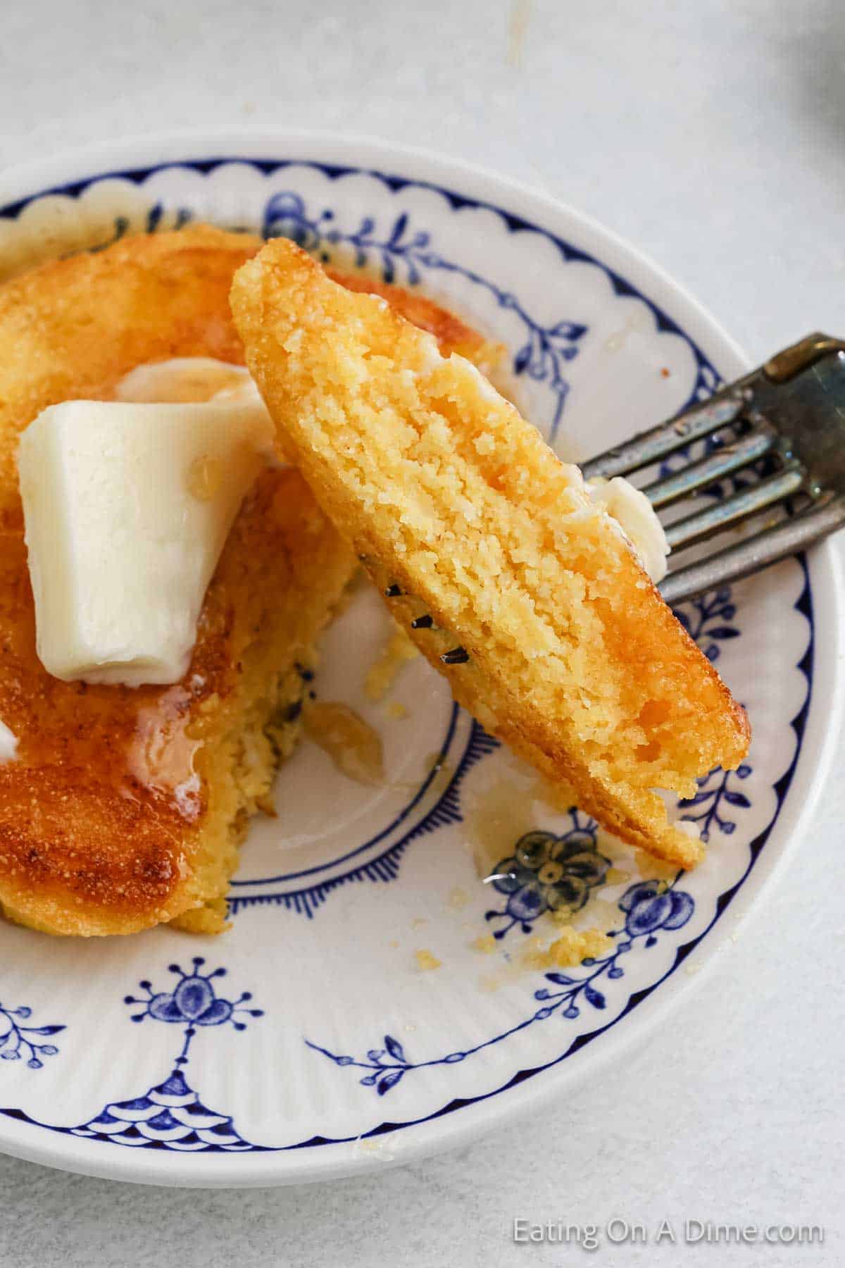 A close-up of a fork holding a piece of fried cornbread with visible crumbs. The cornbread is served on a decorative white plate with blue patterns, topped with a melting pat of butter and drizzled with honey.
