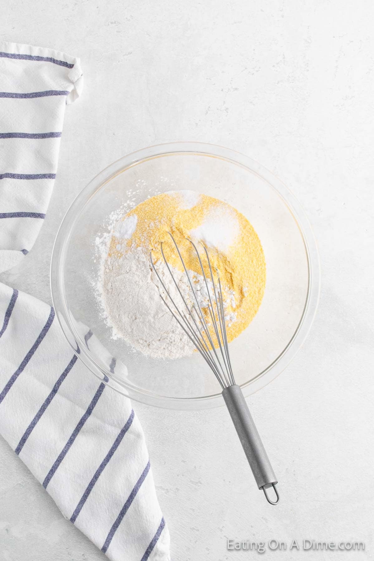 A glass bowl contains a blend of flour, cornmeal, and baking powder—essential components for a delicious fried cornbread recipe. A whisk rests inside, ready to mix. To the left, a white kitchen towel with blue stripes adds a touch of charm to this culinary scene.