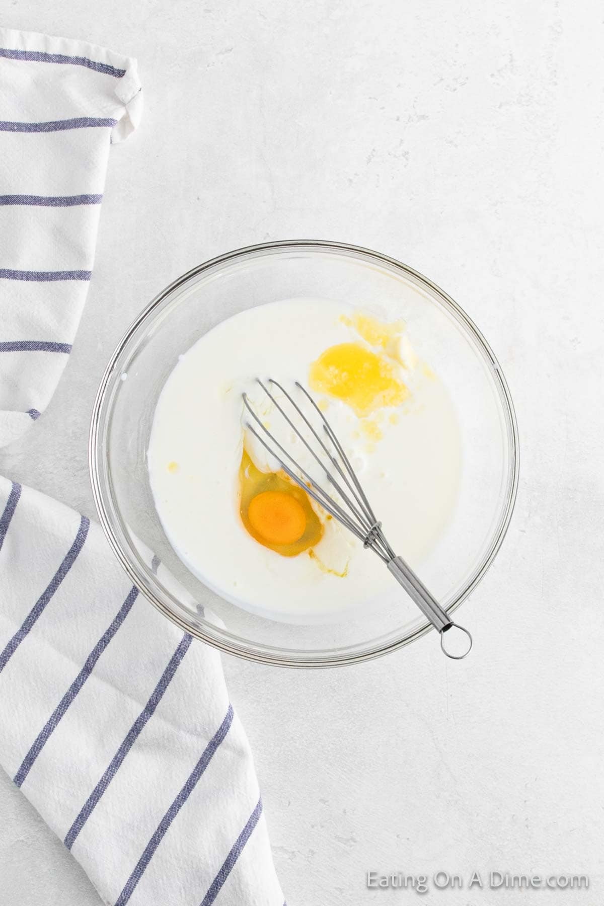 A glass bowl on a white surface contains milk and a cracked egg, with a whisk inside, ready for the fried cornbread recipe. Beside the bowl is a white cloth with navy stripes.