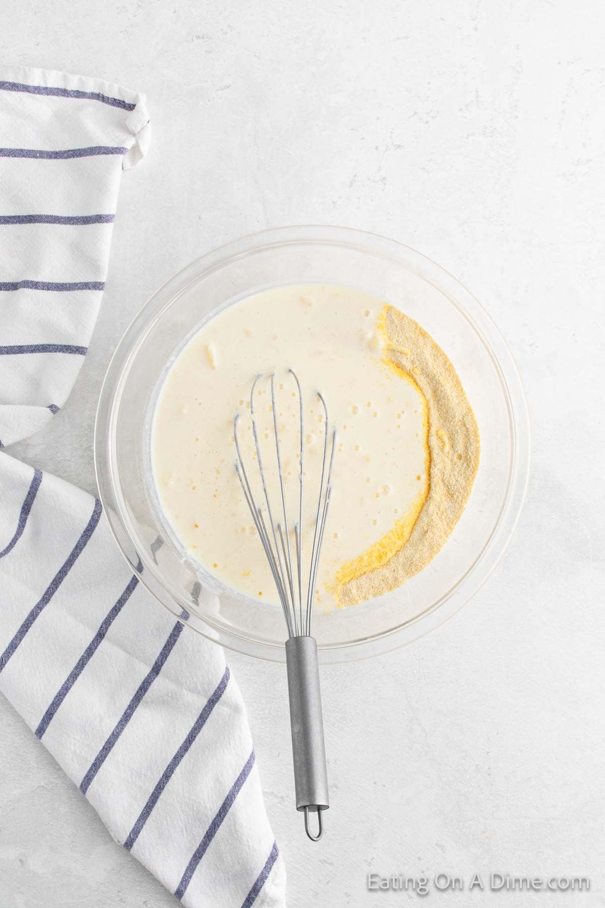 A mixing bowl with cornbread batter and a partial scoop of dry ingredients. A metal whisk is in the bowl, ready to bring the fried cornbread recipe to life, and a striped kitchen towel rests beside it on the white surface.