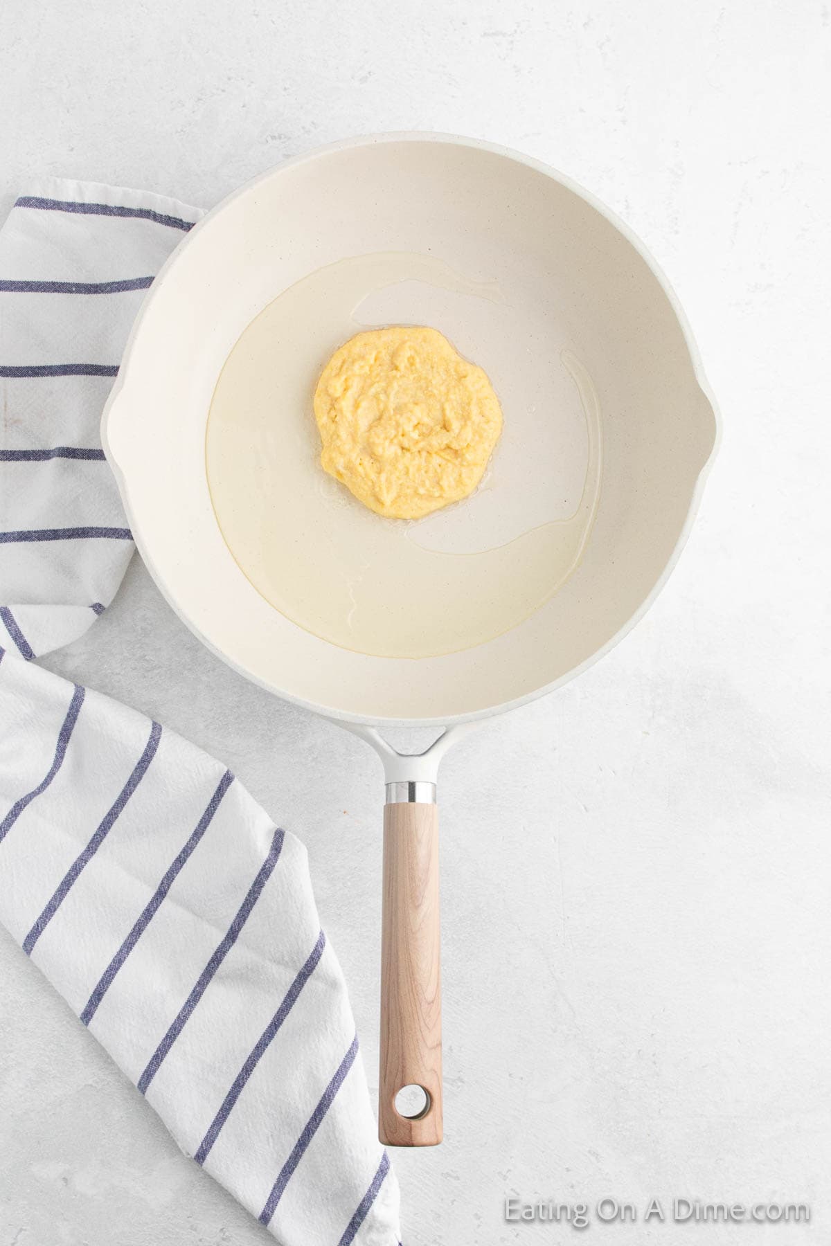 White frying pan with a partially cooked fried cornbread pancake in the center. The pan is on a light gray surface with a white and blue striped kitchen towel beside it, ready to accompany your favorite recipe.