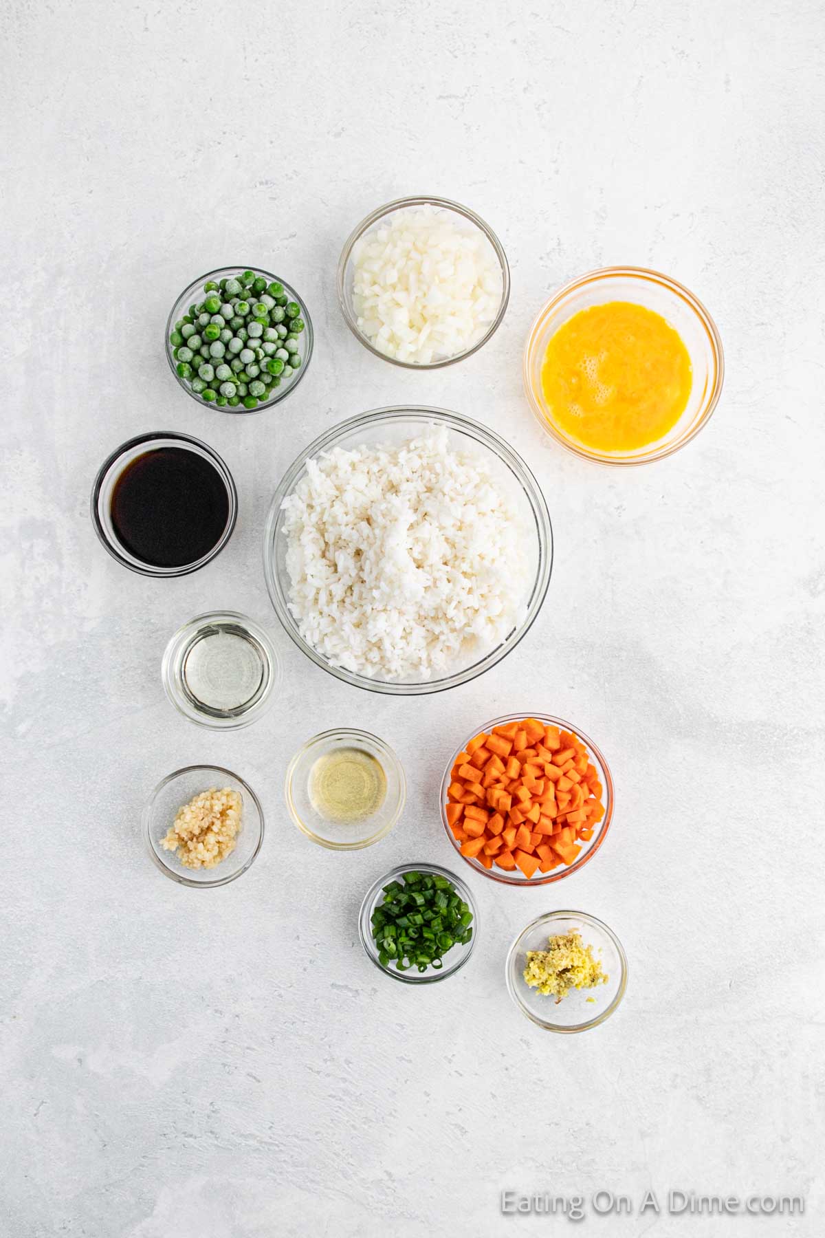 An assortment of ingredients for Hibachi Fried Rice is arranged on a light surface. Central is a bowl of cooked rice, surrounded by peas, chopped carrots, scrambled eggs, garlic, soy sauce, sesame oil, green onions, and ginger.