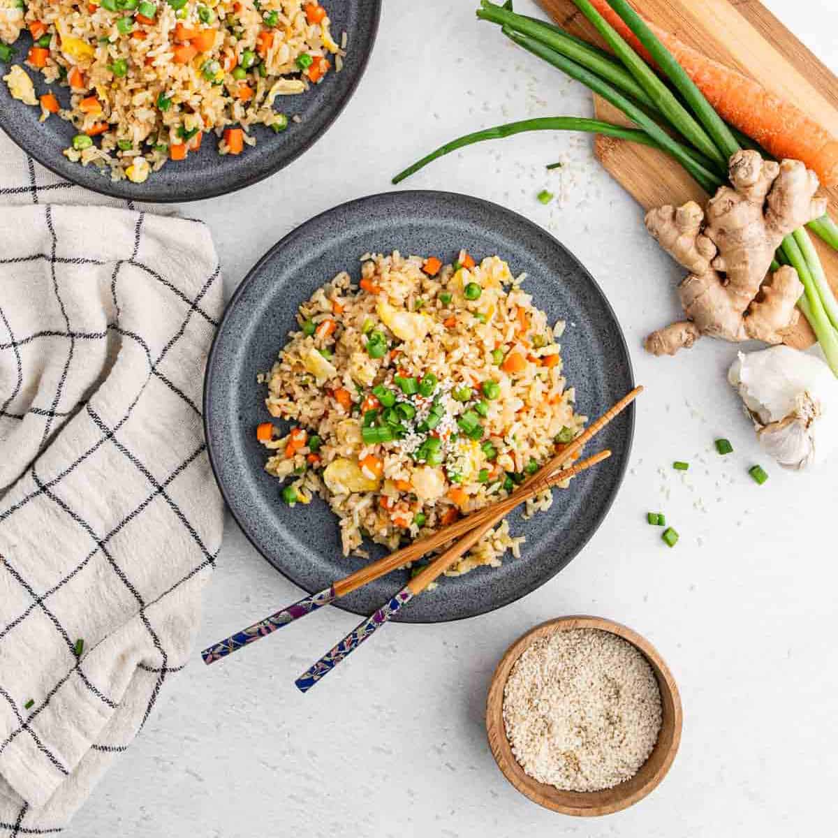 Two plates of hibachi-style fried rice with vegetables, garnished with green onions and accompanied by chopsticks. Fresh ginger, garlic, carrots, and scallions are on a cutting board nearby. A small bowl of sesame seeds and a checkered napkin complete this delicious recipe.