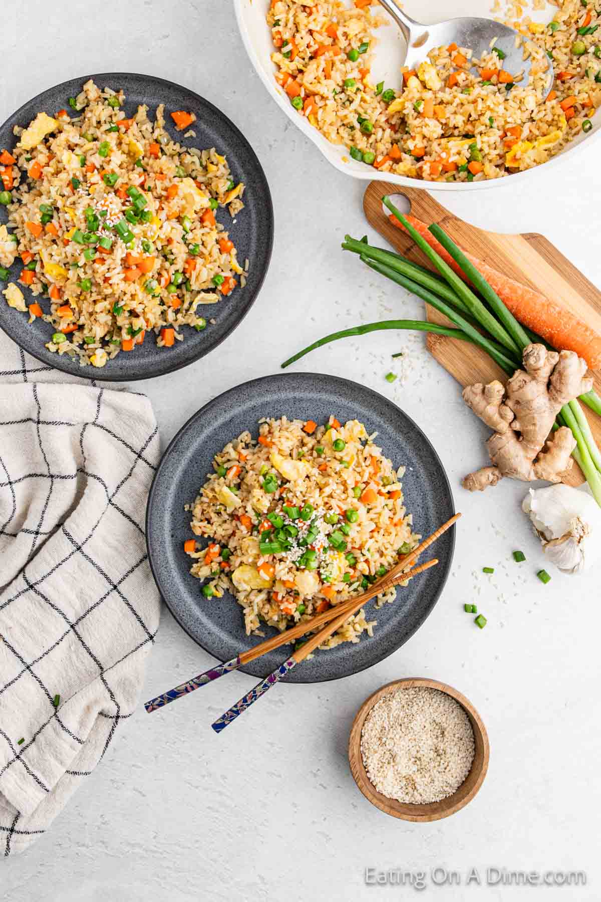 Two plates of hibachi fried rice with vegetables grace a light surface. Fresh ingredients like carrots, green onions, and ginger are nearby. A checkered cloth and a bowl of sesame seeds are also visible. Chopsticks rest on one plate, inviting you to savor this delightful hibachi experience.