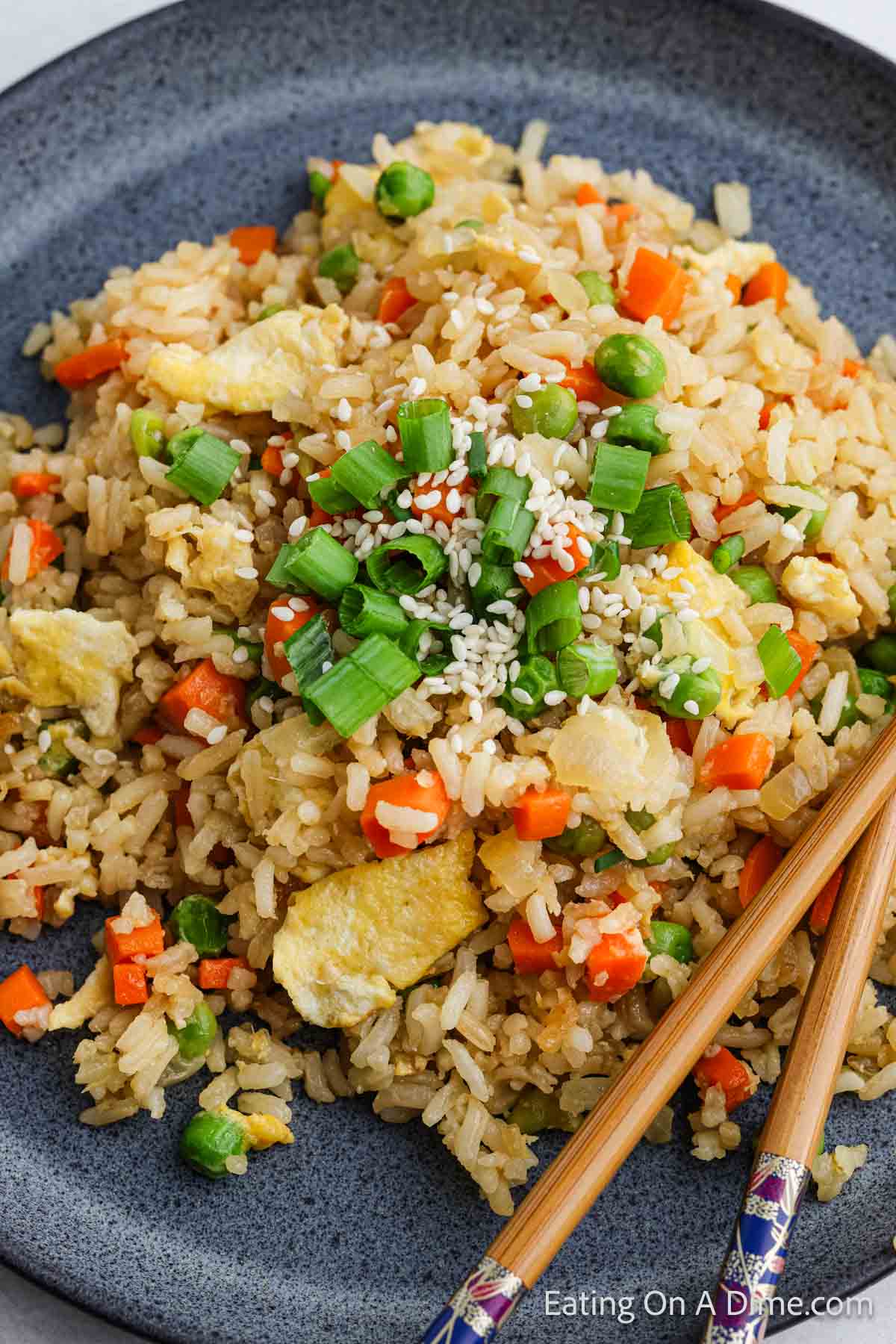 A plate of hibachi fried rice with peas, carrots, scrambled eggs, and green onions. Sesame seeds are sprinkled on top, and a pair of chopsticks is placed on the side.