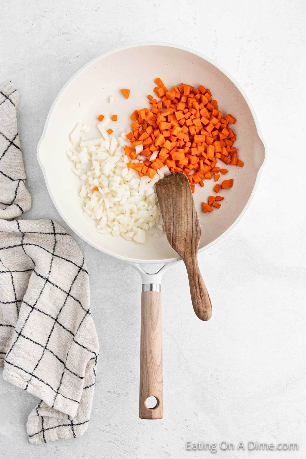 A white skillet on a light surface holds chopped onions and carrots, reminiscent of a hibachi-style fried rice prep, with a wooden spatula resting inside. A checkered cloth is placed beside the skillet.