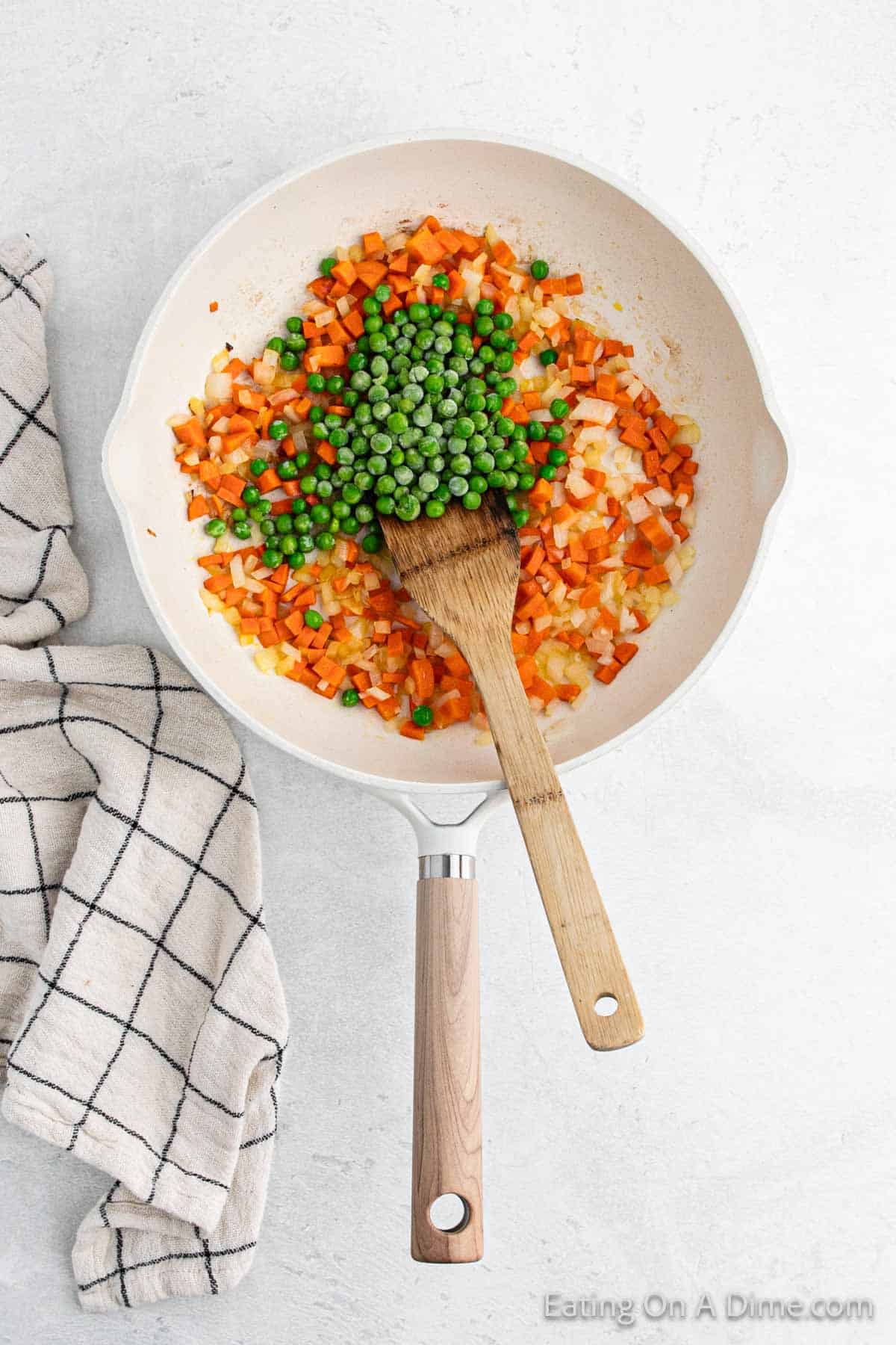 A pan of diced carrots and onions being sautéed like a hibachi star, with a pile of green peas in the center reminiscent of fried rice. A wooden spatula sits in the pan, ready for action, while a checkered cloth rests nearby.