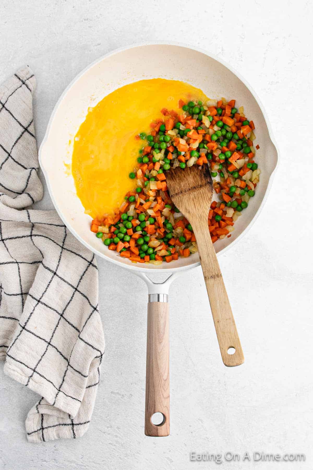 A skillet with scrambled eggs, peas, and diced carrots being stirred with a wooden spatula, reminiscent of a Hibachi fried rice preparation. A checkered kitchen towel rests beside the pan on a white surface.