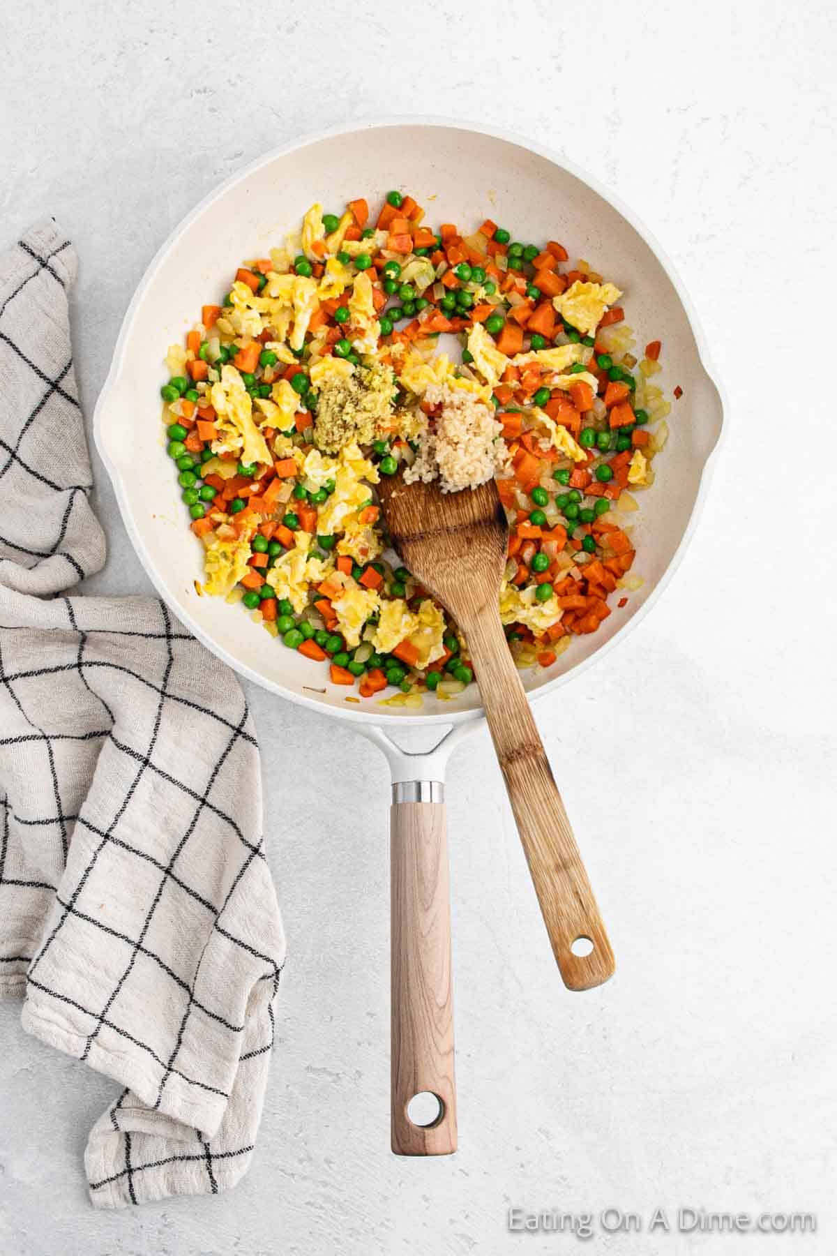 A frying pan brimming with a hibachi-style scramble of eggs, peas, and diced carrots, topped with minced garlic and ginger. A wooden spatula rests inside the pan. A black-and-white checkered kitchen towel lies next to this simple recipe on a light surface.