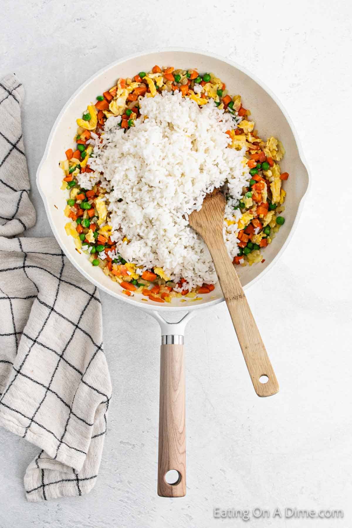 A skillet filled with hibachi-style fried rice, mixed vegetables, and scrambled eggs rests on a light surface. A wooden spoon is in the skillet, and a checkered kitchen towel is laid beside it.