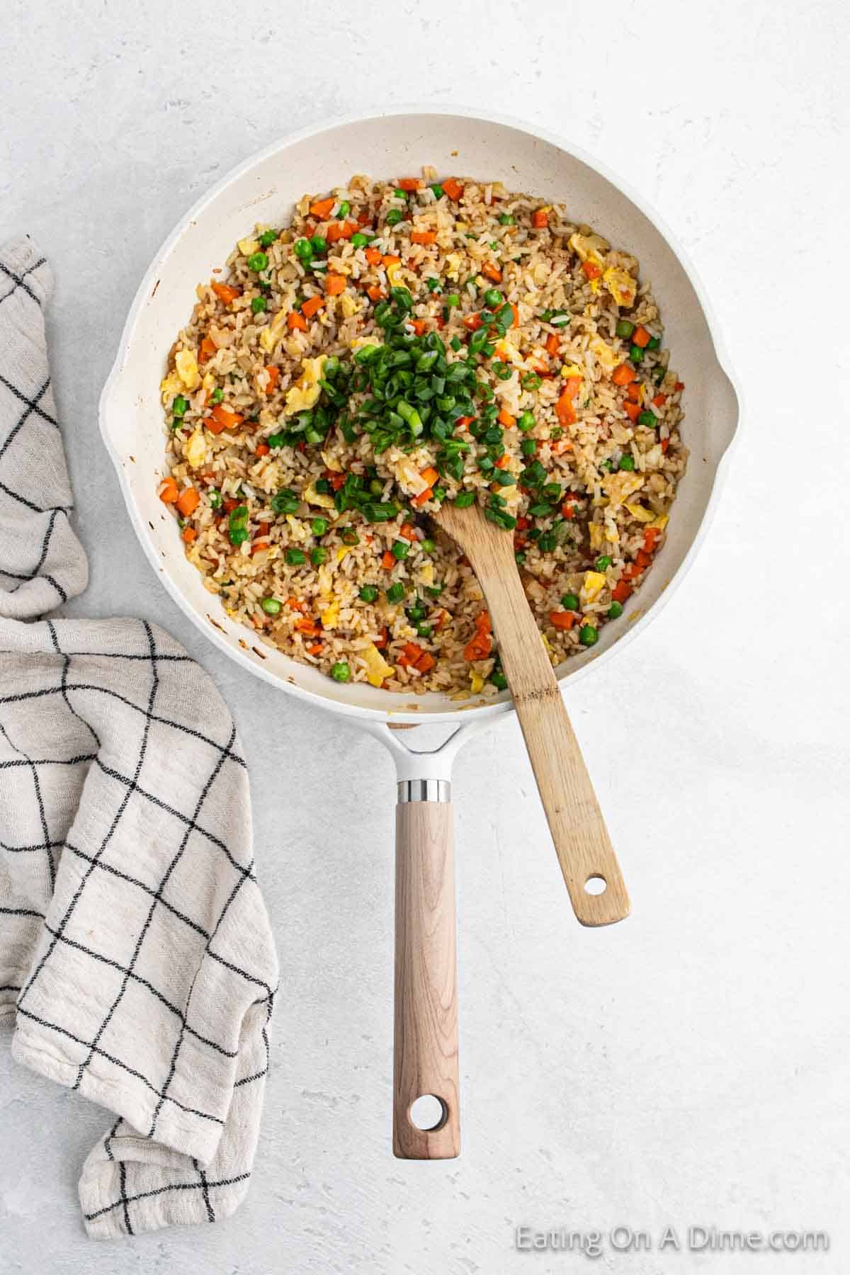 A skillet brimming with vibrant hibachi fried rice, featuring peas, carrots, and scrambled eggs. A wooden spoon rests inside, garnished with chopped green onions. A folded checkered cloth lies beside the skillet on a light-colored surface.