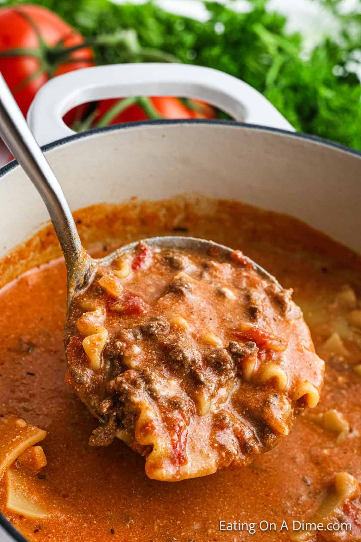 A close-up of a hearty lasagna soup in a pot, with a ladle scooping a portion. This comforting recipe is rich and creamy, featuring visible pasta, ground meat, and tomatoes. Fresh greens and extra tomatoes add a burst of color in the background.