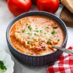 A bowl of creamy lasagna-inspired soup with pasta and ground meat, topped with shredded cheese and fresh herbs. A spoon rests in the bowl, surrounded by fresh tomatoes and a slice of bread. A checkered cloth is nearby for a cozy touch.