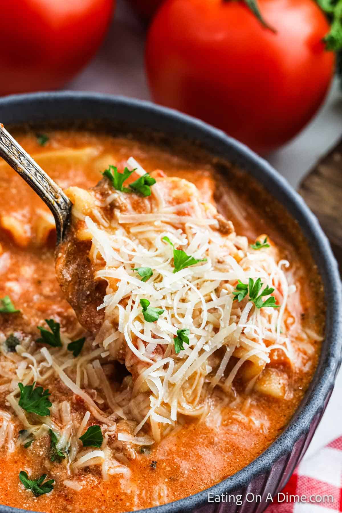 A bowl of creamy tomato lasagna soup with shredded cheese and parsley on top. A spoon lifts a portion, revealing its rich texture. Fresh tomatoes are visible in the background, inviting you to savor every bite.