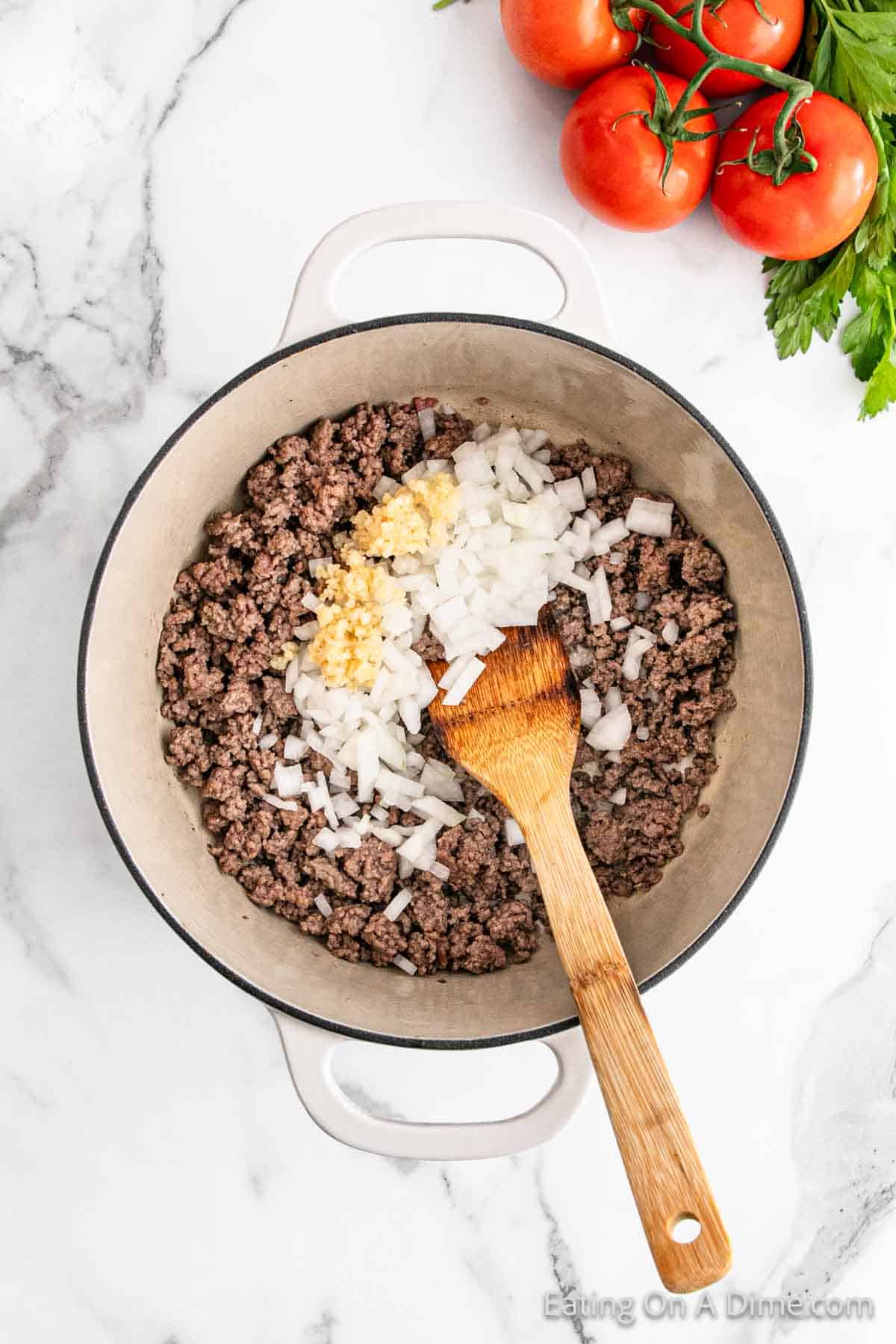 A white pot on a marble surface holds cooked ground beef layered with chopped onions and minced garlic, reminiscent of a savory lasagna. A wooden spoon rests within the rich mixture. Nearby, fresh tomatoes and parsley await to enhance the dish's vibrant flavors.