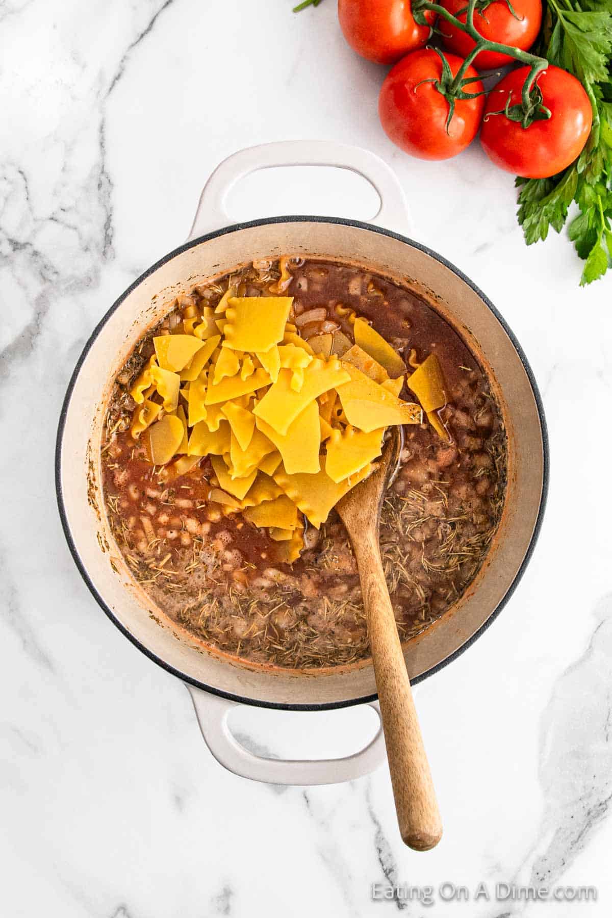 A white pot on a marble counter holds a delicious lasagna soup, with chunks of orange cheese and a wooden spoon. Fresh tomatoes and herbs are artfully arranged beside the pot, inviting you to try this comforting recipe.