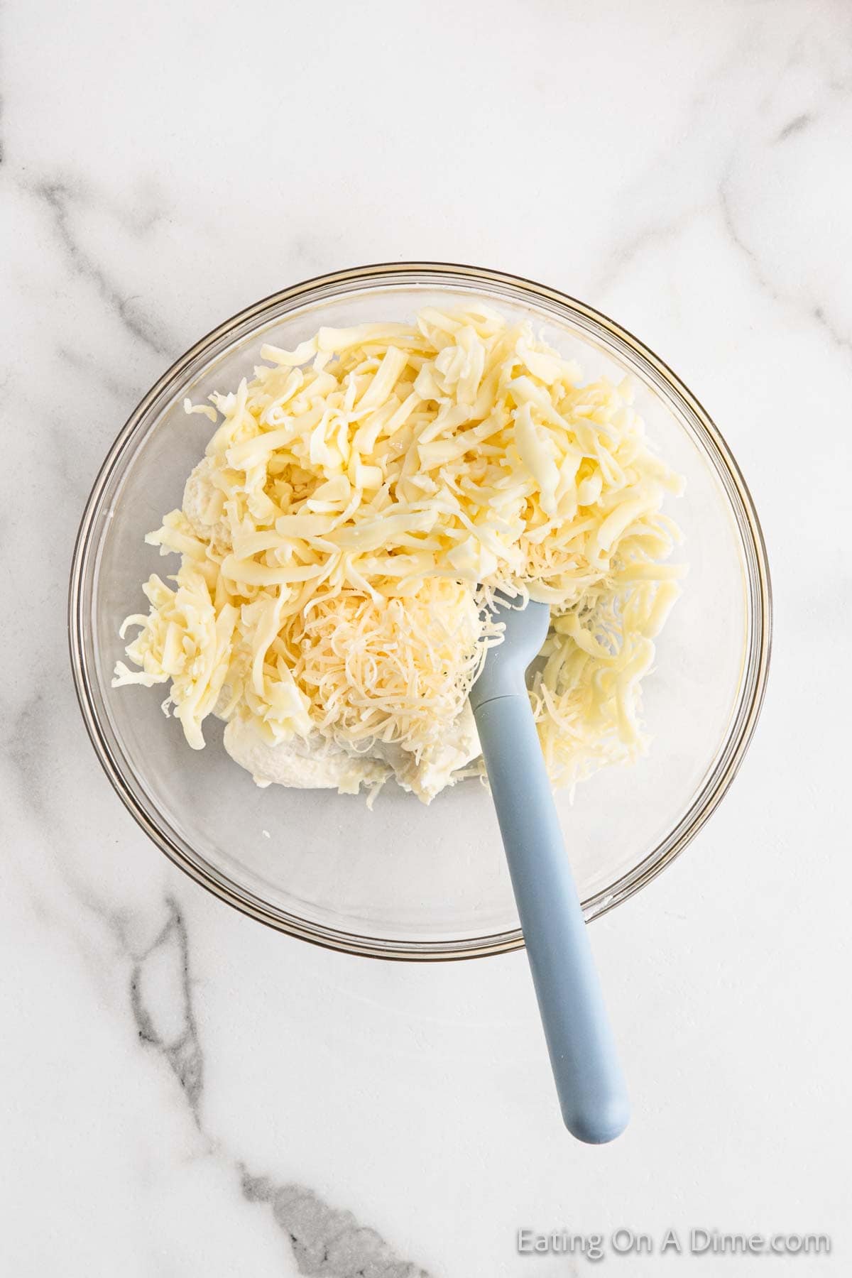 A clear glass bowl filled with shredded cheese, essential for a savory lasagna soup, with a blue spatula resting inside. The bowl is placed on a white marble surface.