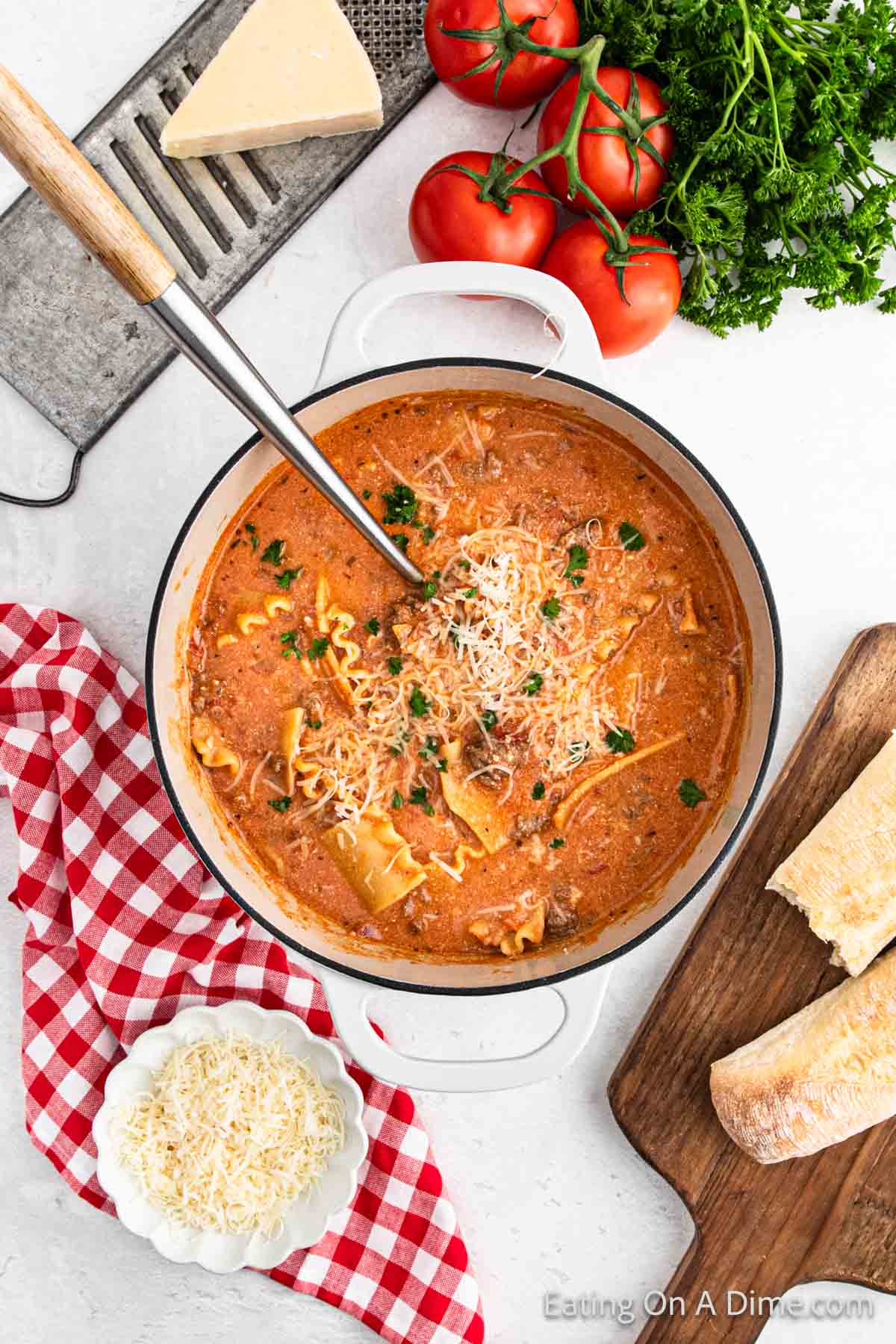 A pot of creamy tomato lasagna soup topped with grated cheese and parsley. Surrounding the pot are fresh tomatoes, parsley, and a wedge of cheese. Nearby, a bowl of shredded cheese, a grater, and sliced bread rest on a wooden board.