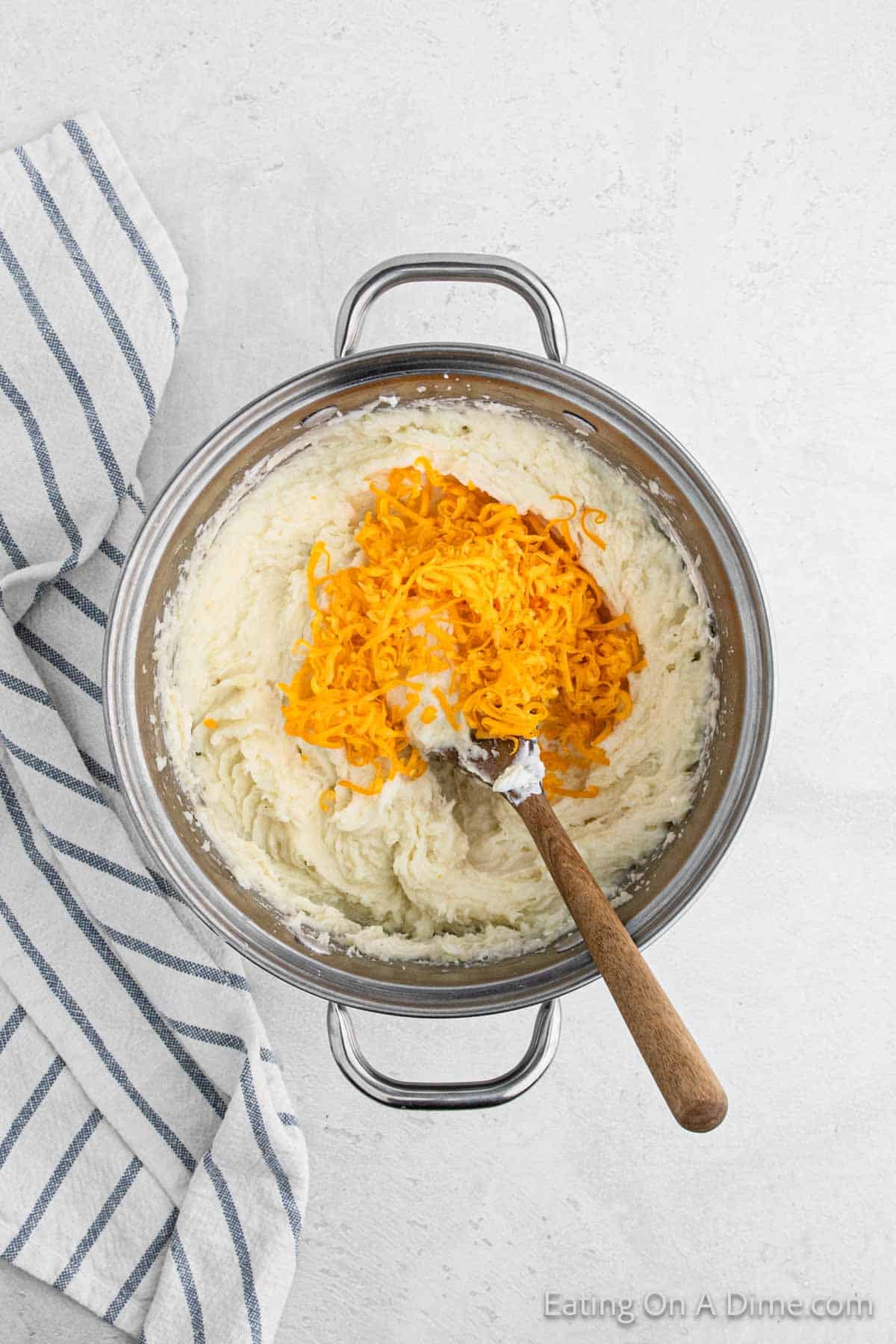 Mashed potatoes in a large pot topped with shredded cheese with a wooden spoon