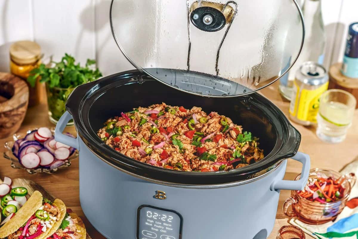 A slow cooker filled with a colorful dish of shredded meat, vegetables, and herbs sits on a wooden countertop. With sliced radishes, fresh cilantro, tacos, a drink can, and glassware around it, this vibrant food scene is perfect for those preparing festive meals inspired by Early Black Friday Deals.